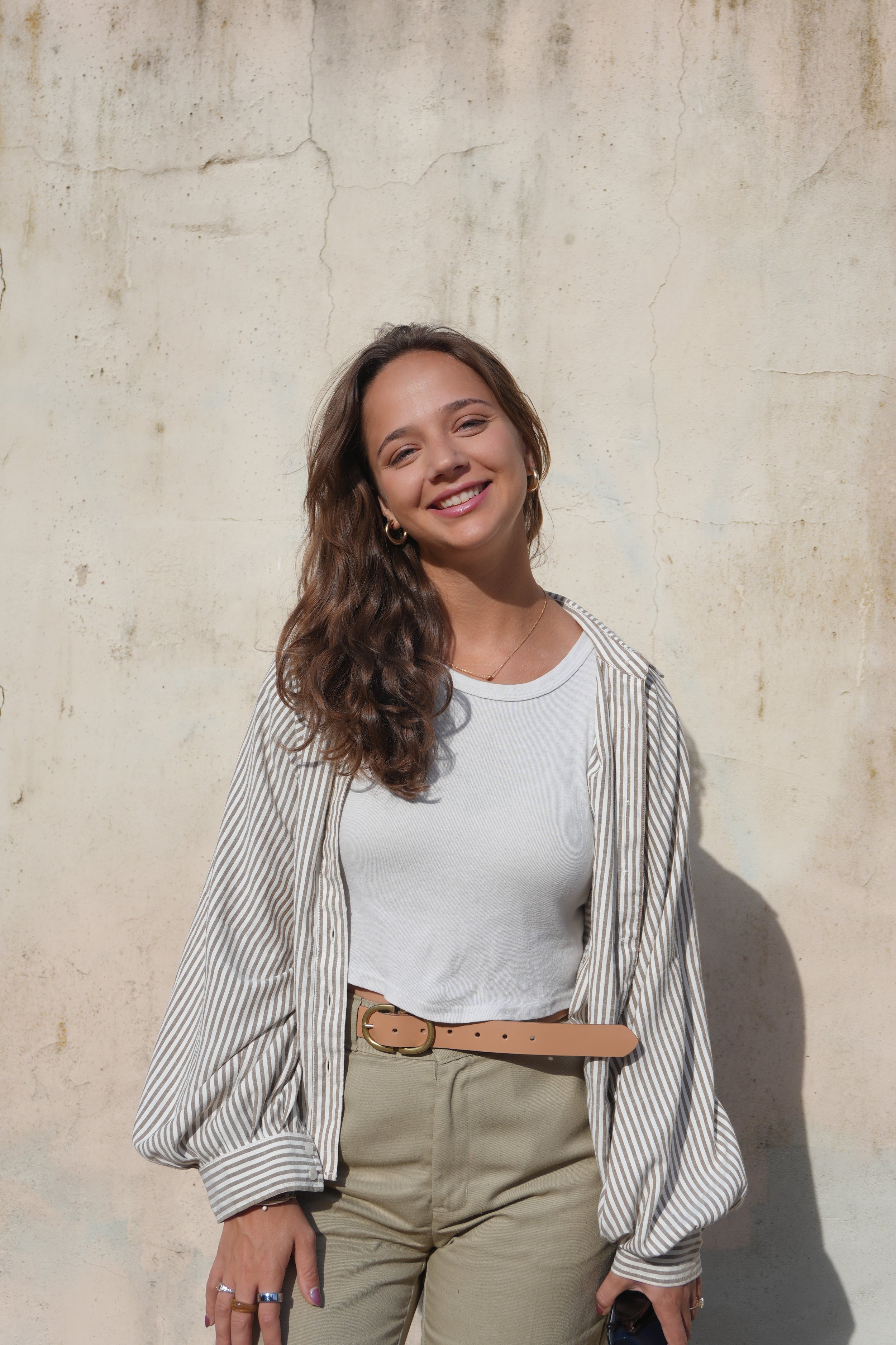 Young woman with long curly hair, smiling, wearing a white T-shirt, beige pants, tan belt, and a striped shirt draped over her shoulders, standing against a weathered beige wall.