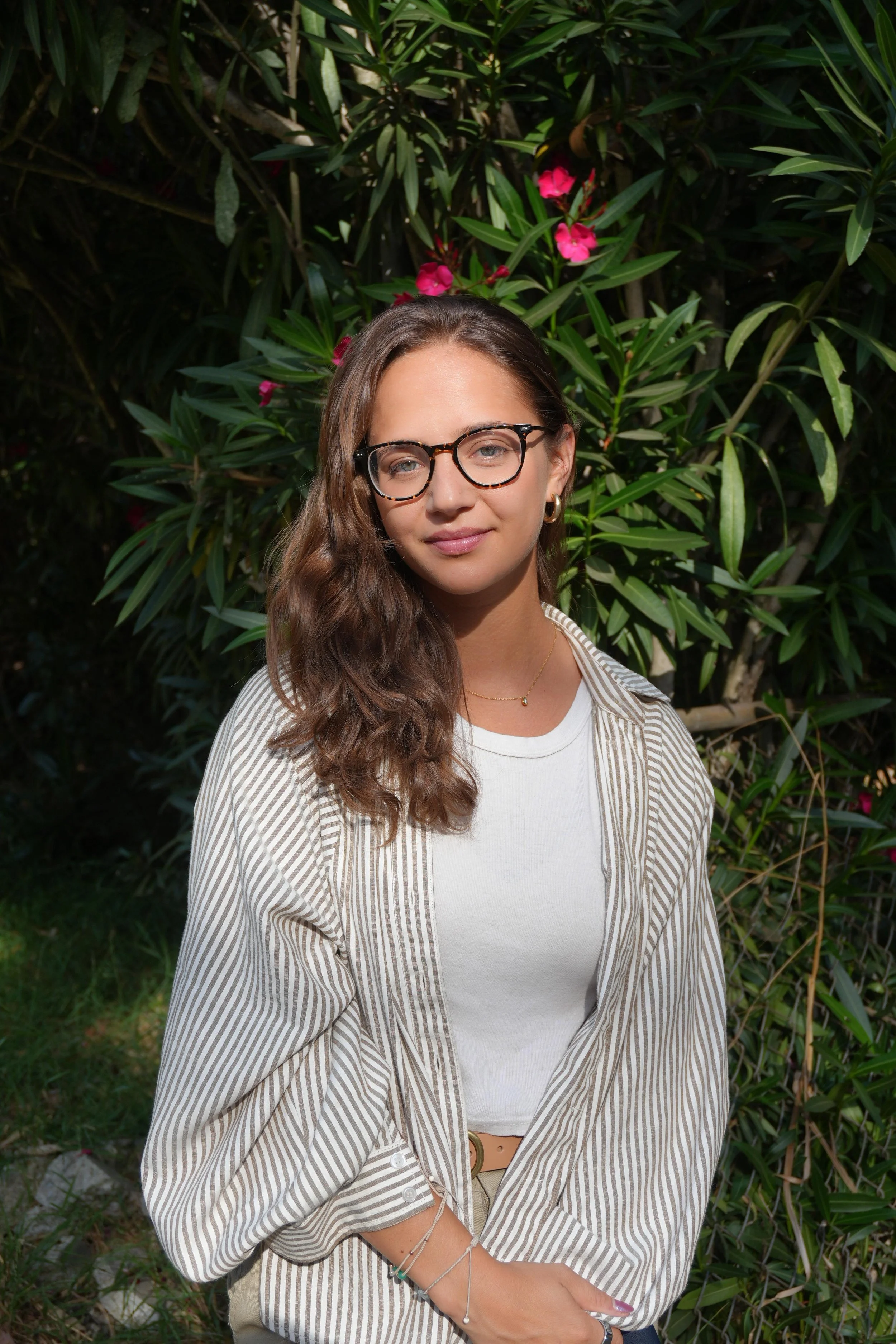 A young woman with long wavy brown hair, wearing glasses, a white top, and a beige and white striped button-up shirt, stands outdoors in front of green leafy bushes with some pink flowers. She has gold hoop earrings, a delicate necklace, and multiple bracelets on her left wrist, with her arms crossed.