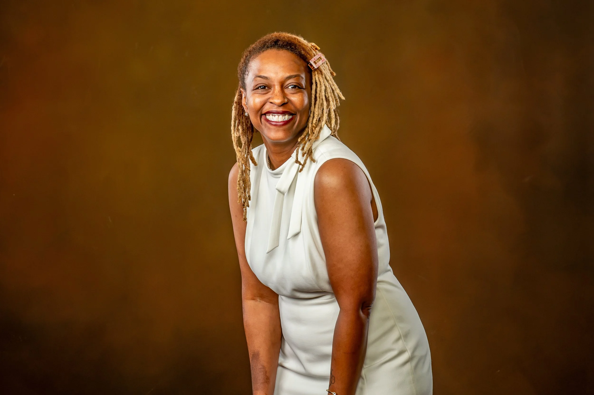 Smiling woman with dreadlocks, wearing a white sleeveless dress, standing against a brown background.