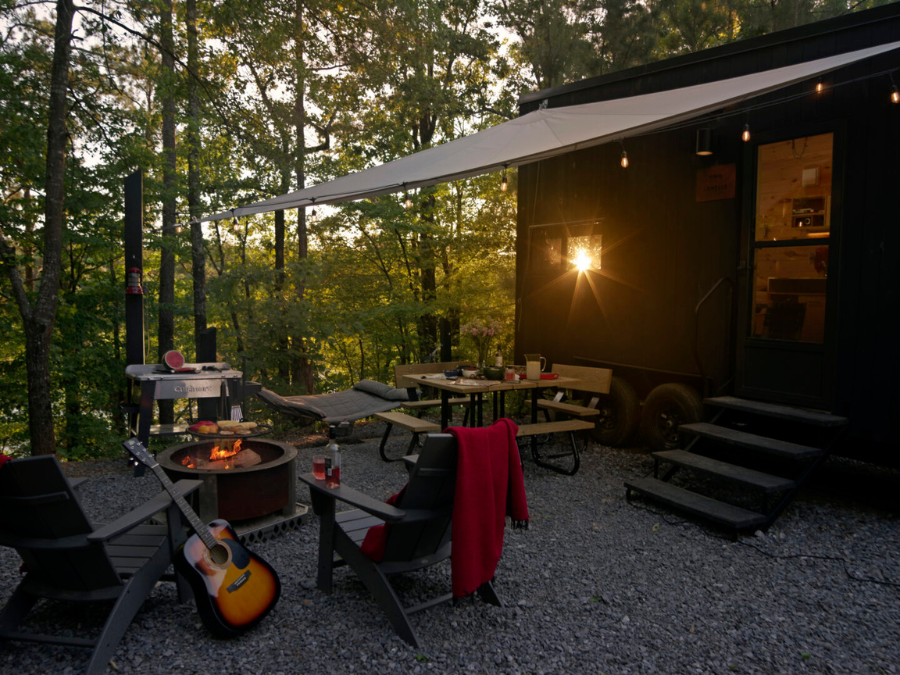 A small black trailer cabin next to a campfire set up with a guitar and two chairs