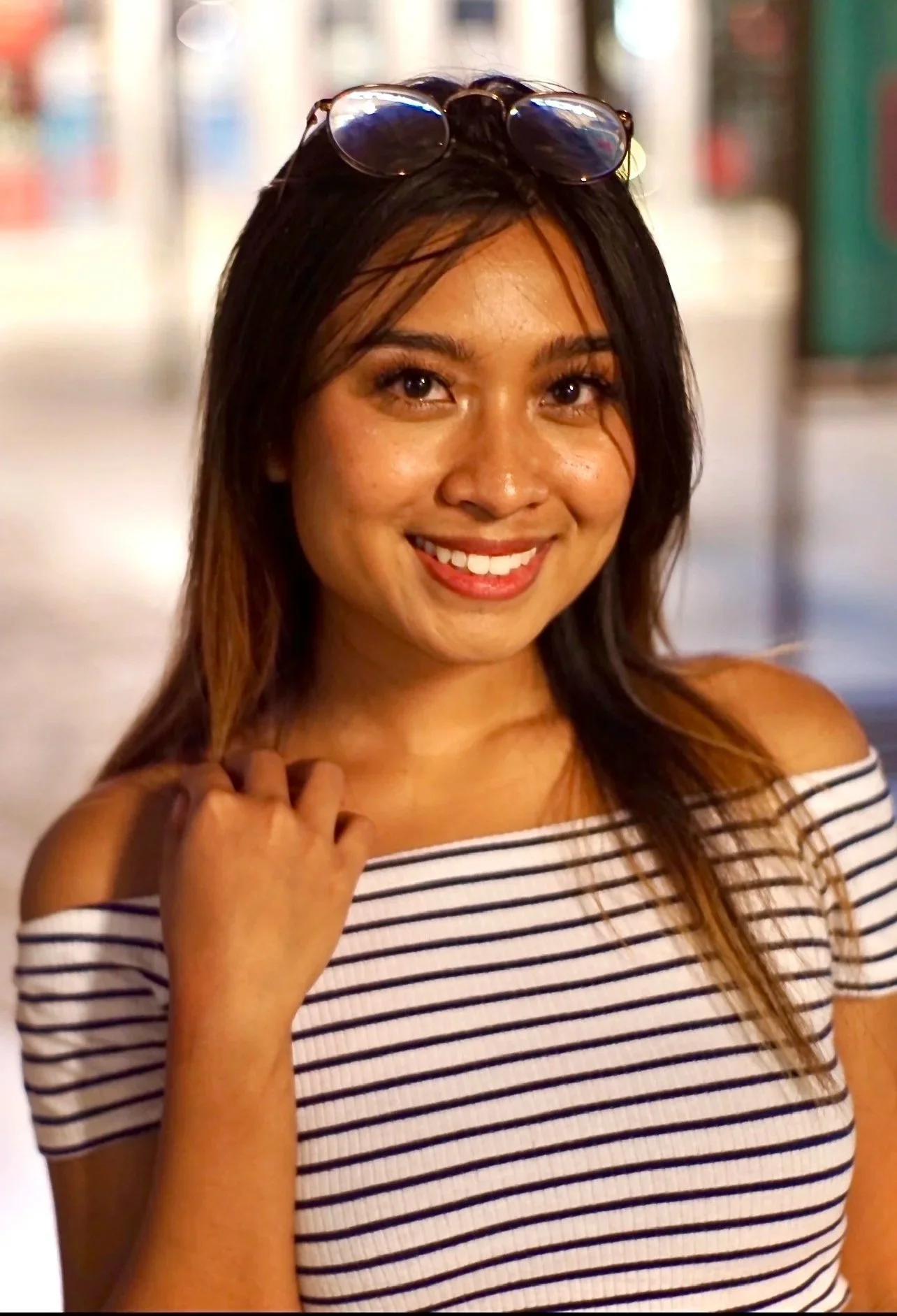 A woman smiling, wearing a striped off-shoulder top and sunglasses on her head, with a background of bookshelves.