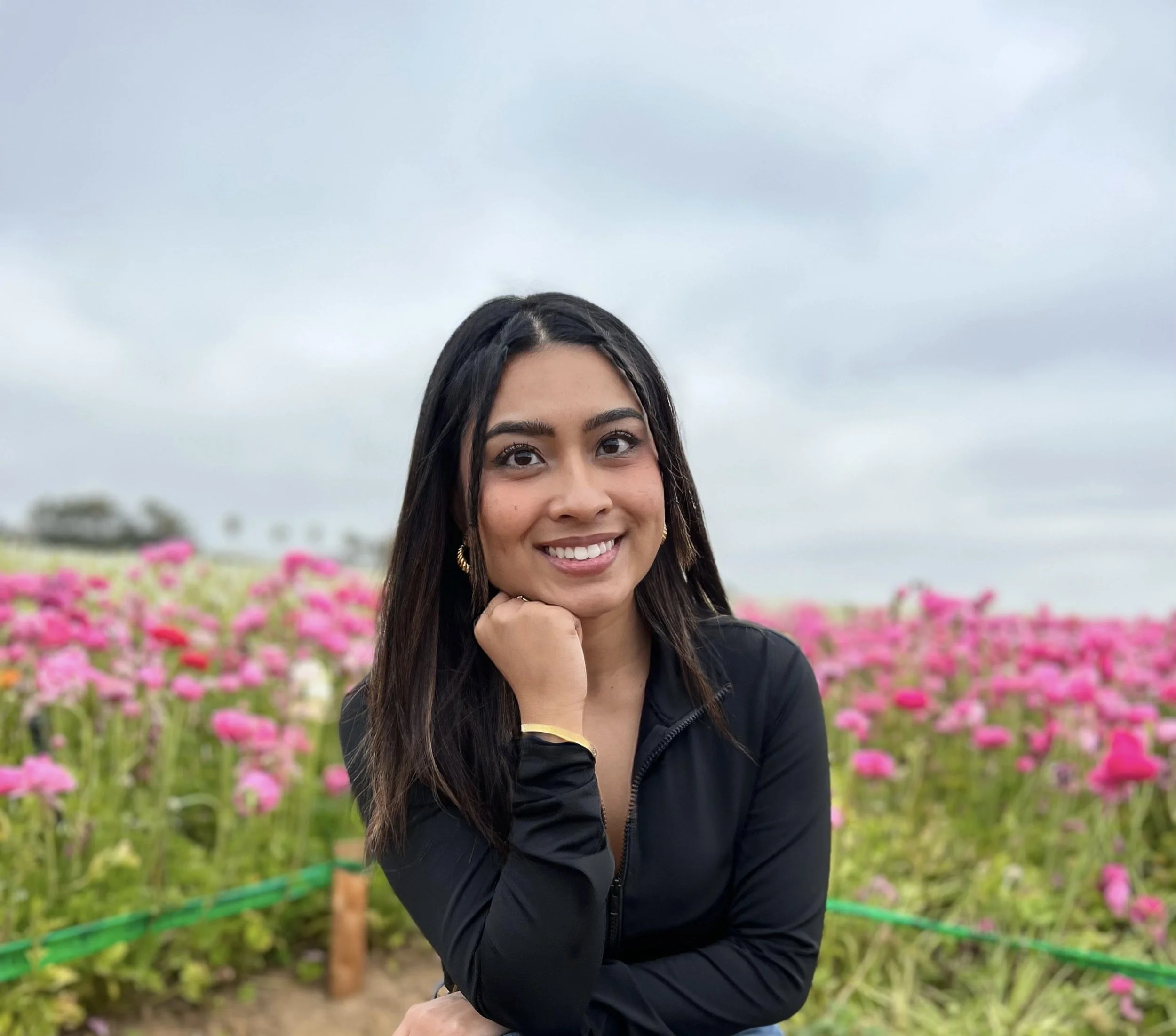 A woman with long dark hair smiling in a flower field with pink blossoms.