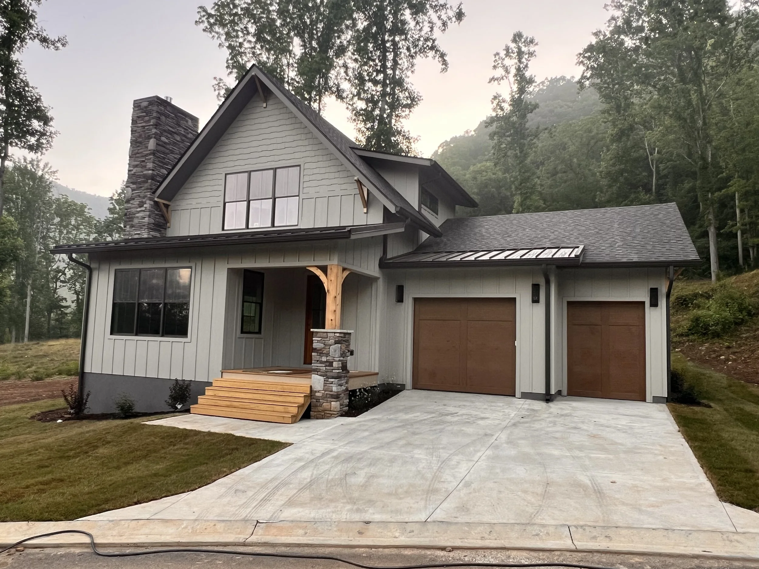 Front view of a new two-story house with a stone chimney, white siding, large windows, a covered porch with wood steps, and attached garage, surrounded by trees.