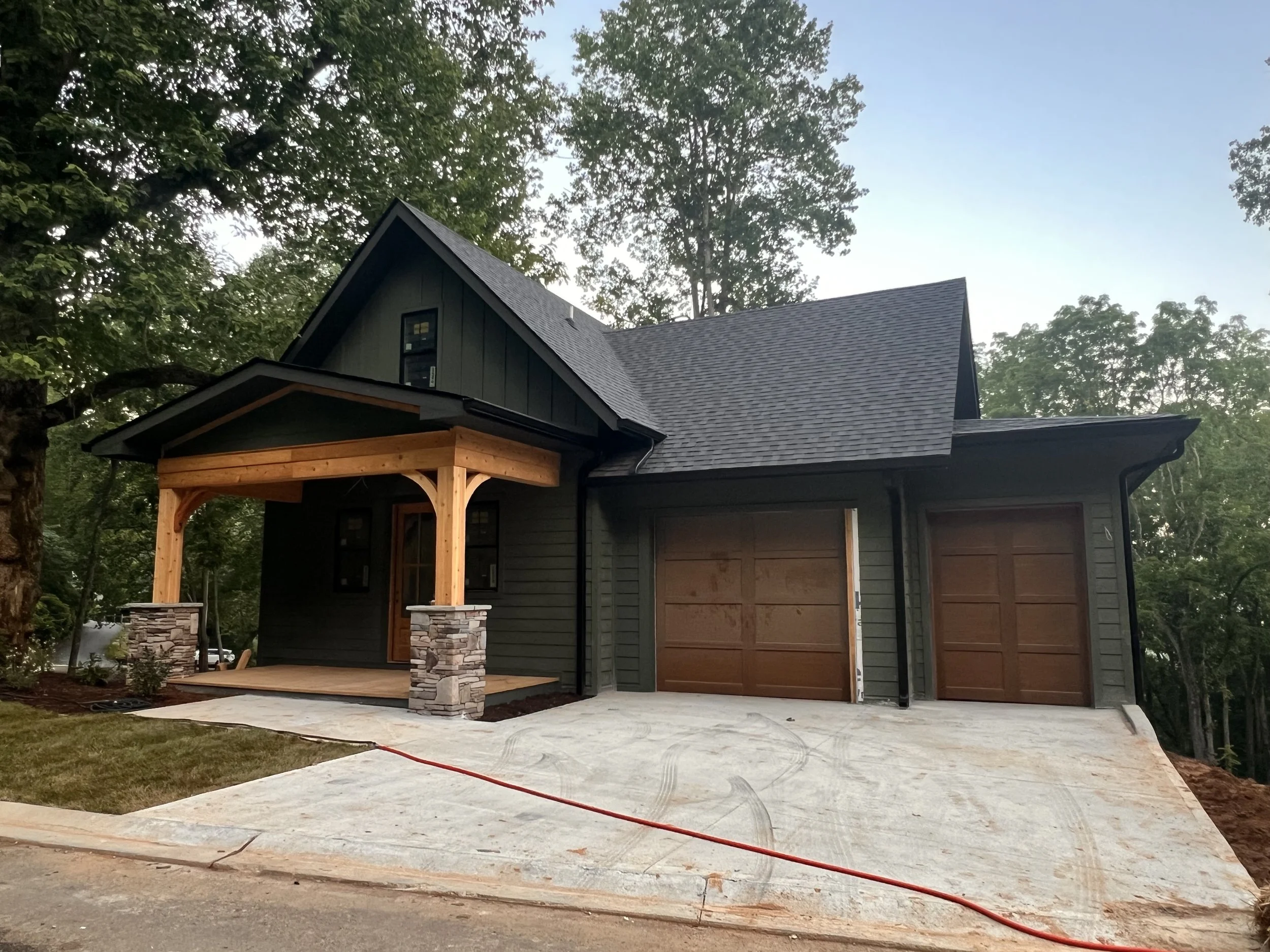 A house under construction with a grey roof, dark green siding, a wooden porch with stone columns, and a concrete driveway, surrounded by trees.
