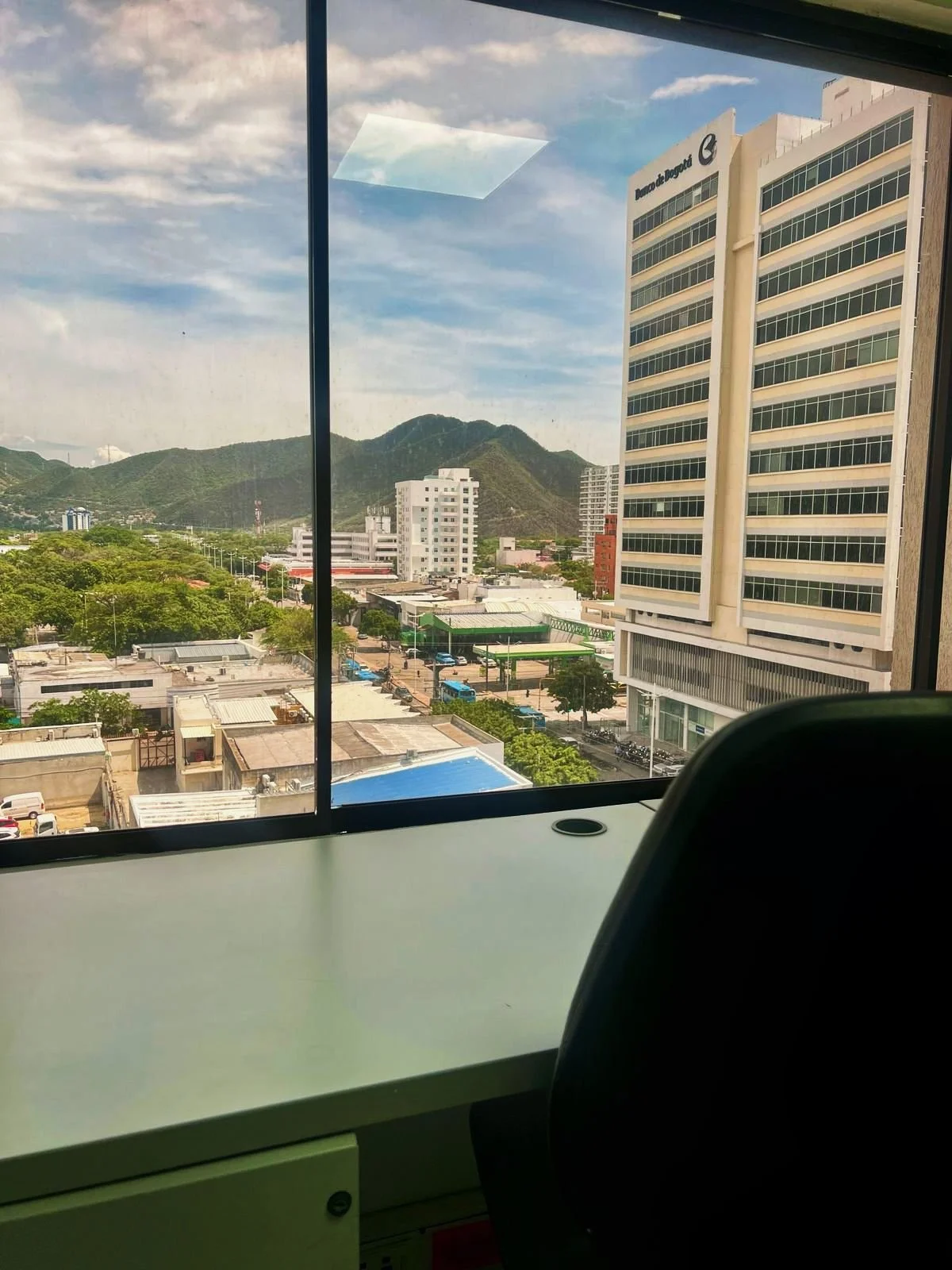 Vista desde una ventana de un edificio, con vista a una ciudad con edificios altos, áreas verdes y montañas en el fondo, y un cielo parcialmente nublado.