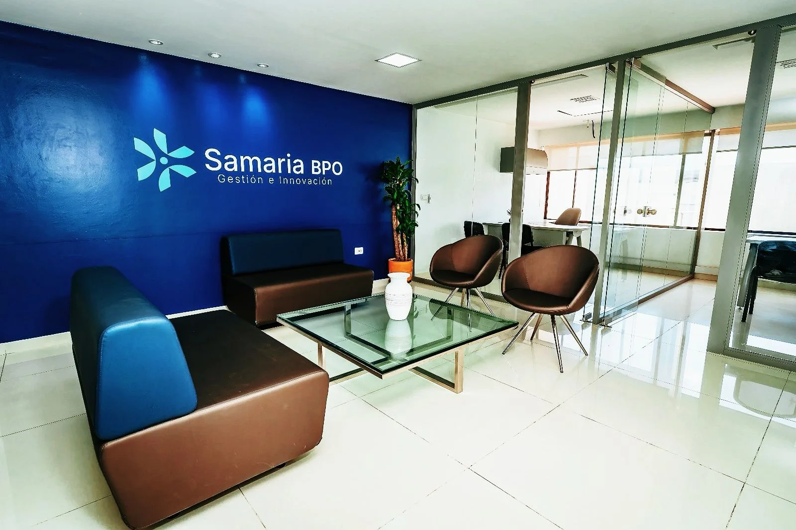 Sala de espera moderna con sillones de cuero, mesa de vidrio y un muro azul con el logo y nombre de 'Samaria BPO Gestión e Innovación'.
