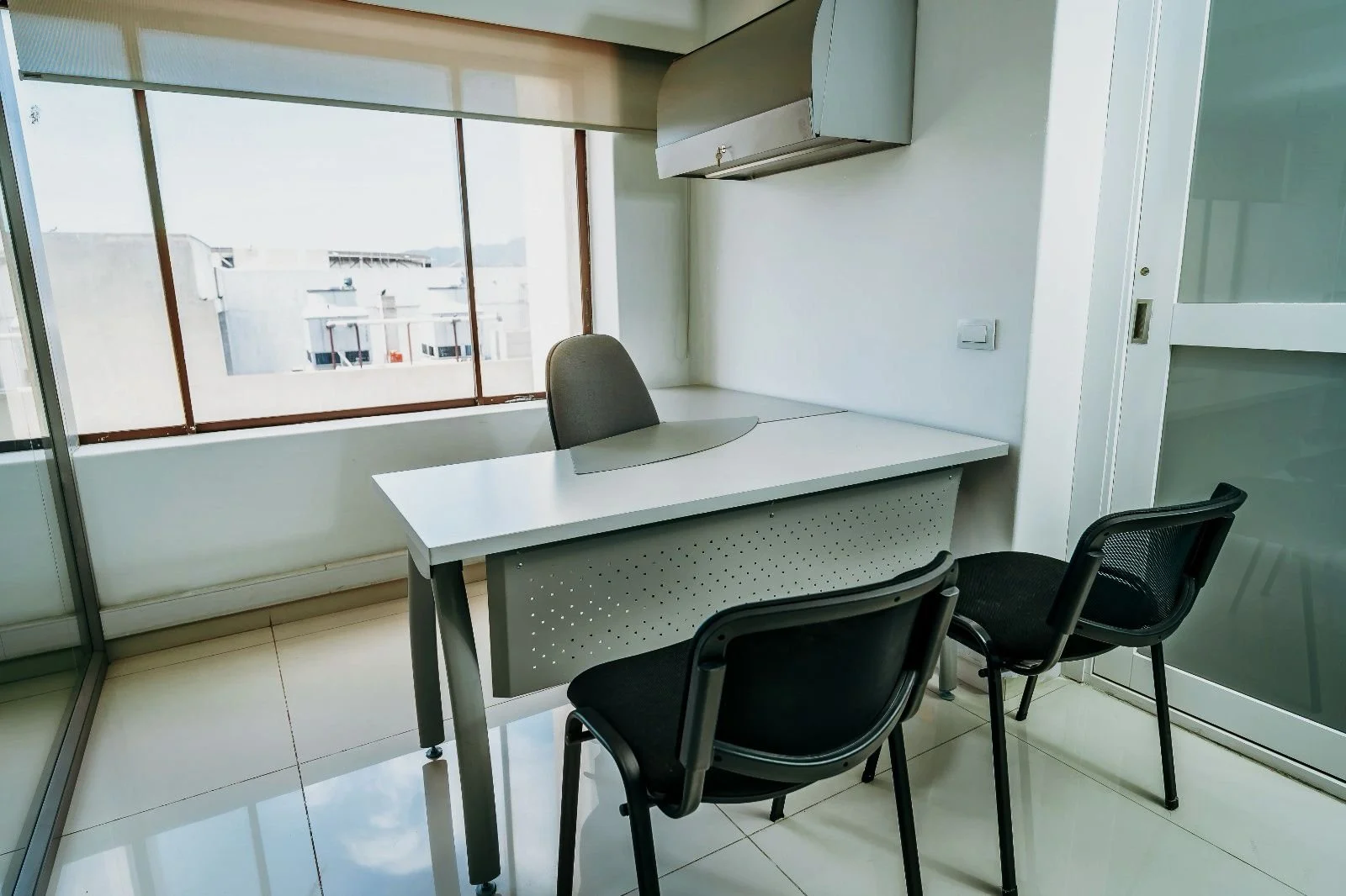 Espacio de oficina con una mesa blanca, tres sillas negras, y ventanas grandes que dejan pasar mucha luz natural.