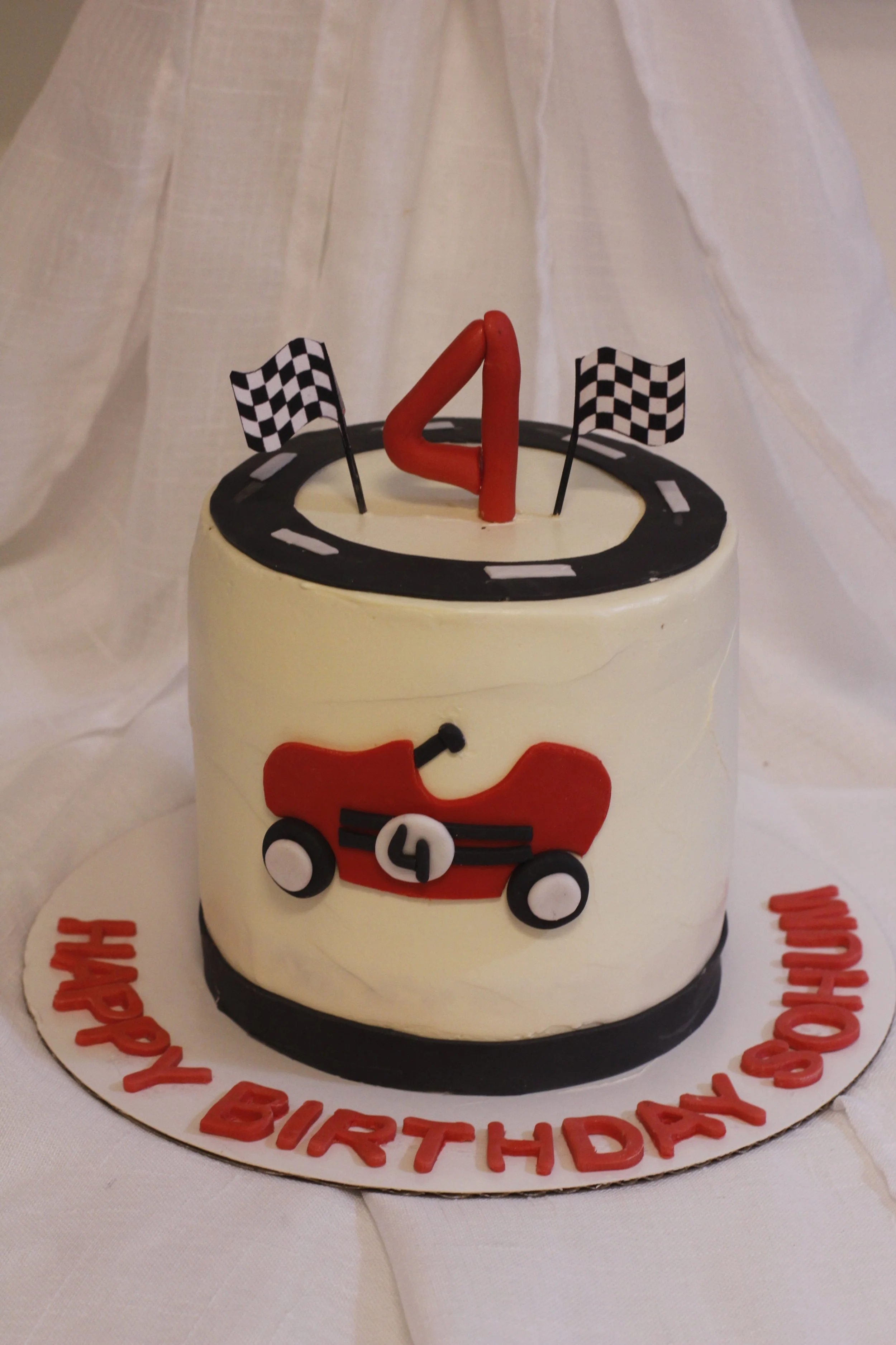 A birthday cake decorated with a racing theme, featuring black and white checkered flags, a red number 4 candle, a race car design, and the message "Happy Birthday Soham" on the base.