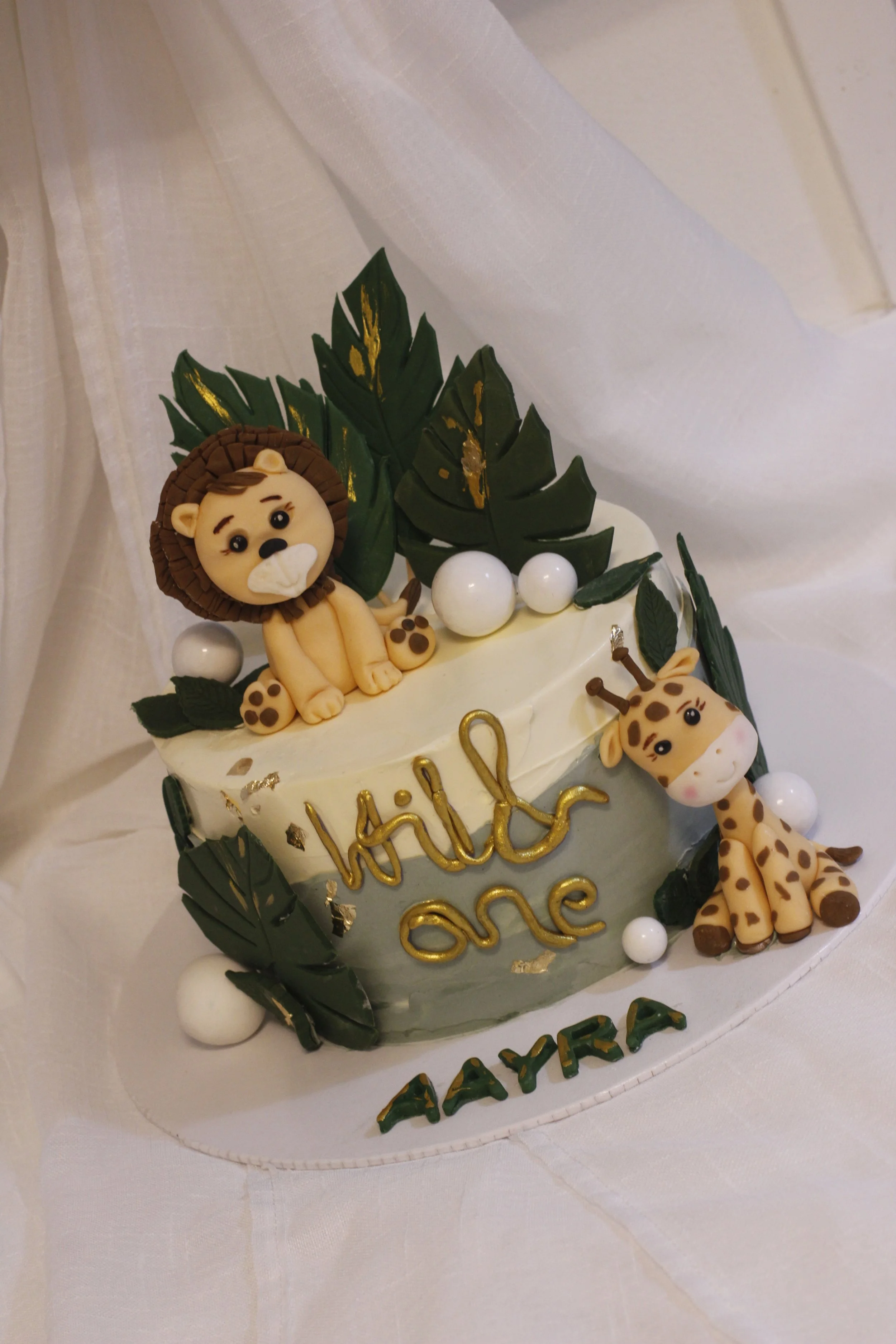A decorated cake with jungle animals, including a lion and a giraffe, surrounded by green leaves, white decorative balls, and a gold "hello one" sign, with the name 'Aayra' written at the bottom.