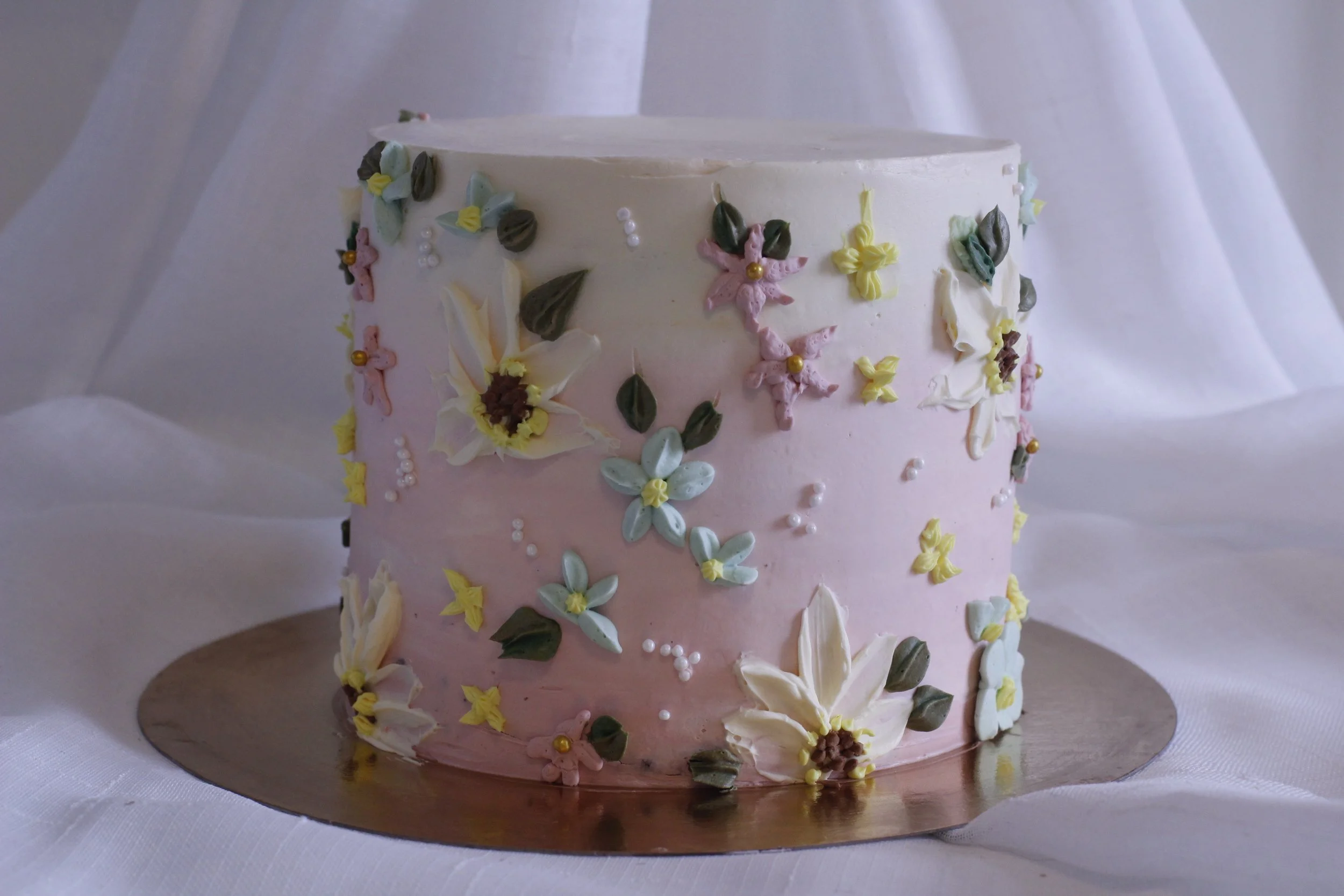 A round wedding cake with pink and white buttercream icing, decorated with intricate pastel-colored floral piping and small pearl-like accents, set on a silver cake board with white fabric backdrop.