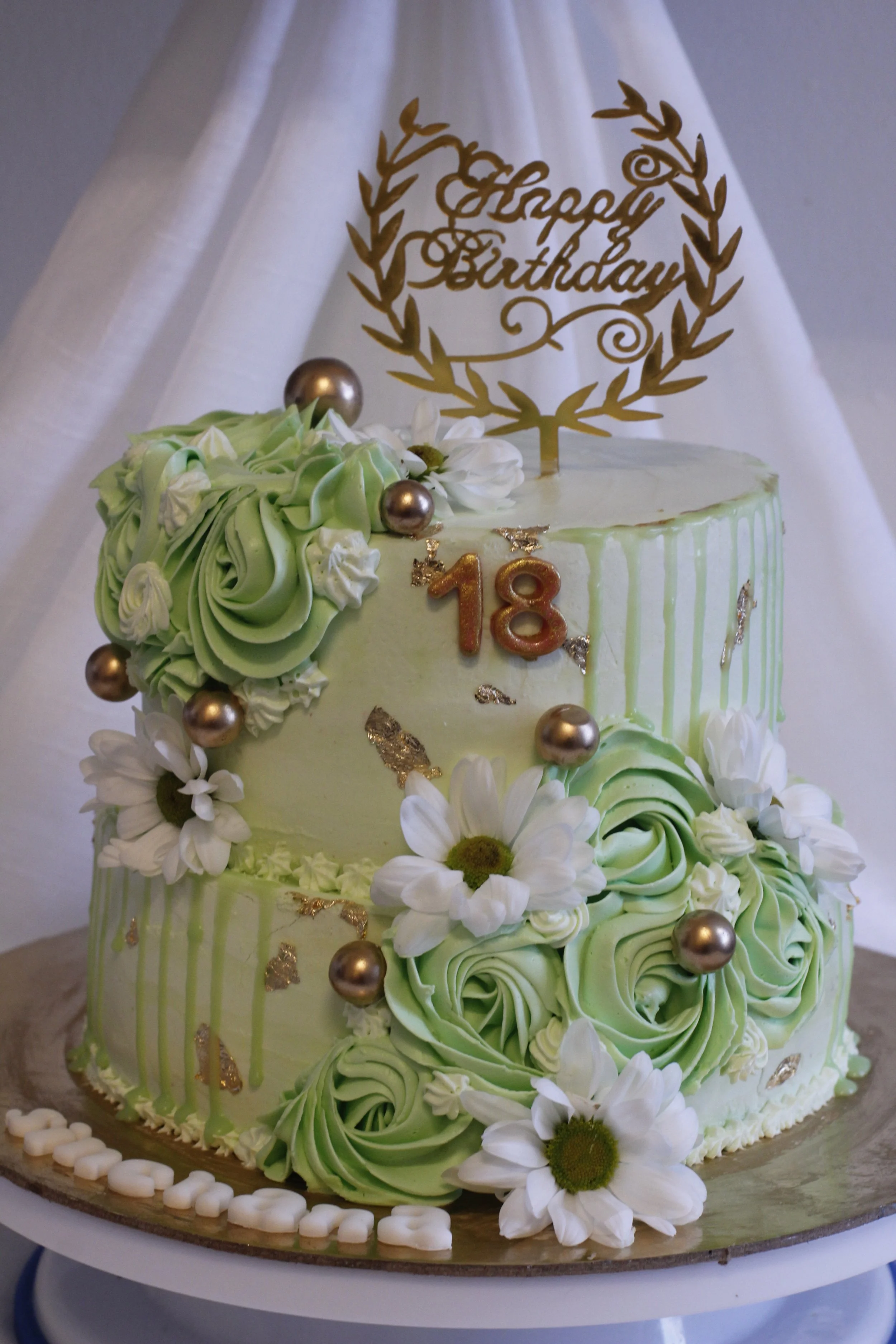 Two-tiered birthday cake decorated with white and green swirls, white daisies, gold accents, and a gold "Happy Birthday" topper. The cake has the number "18" on it and is placed on a silver cake board.