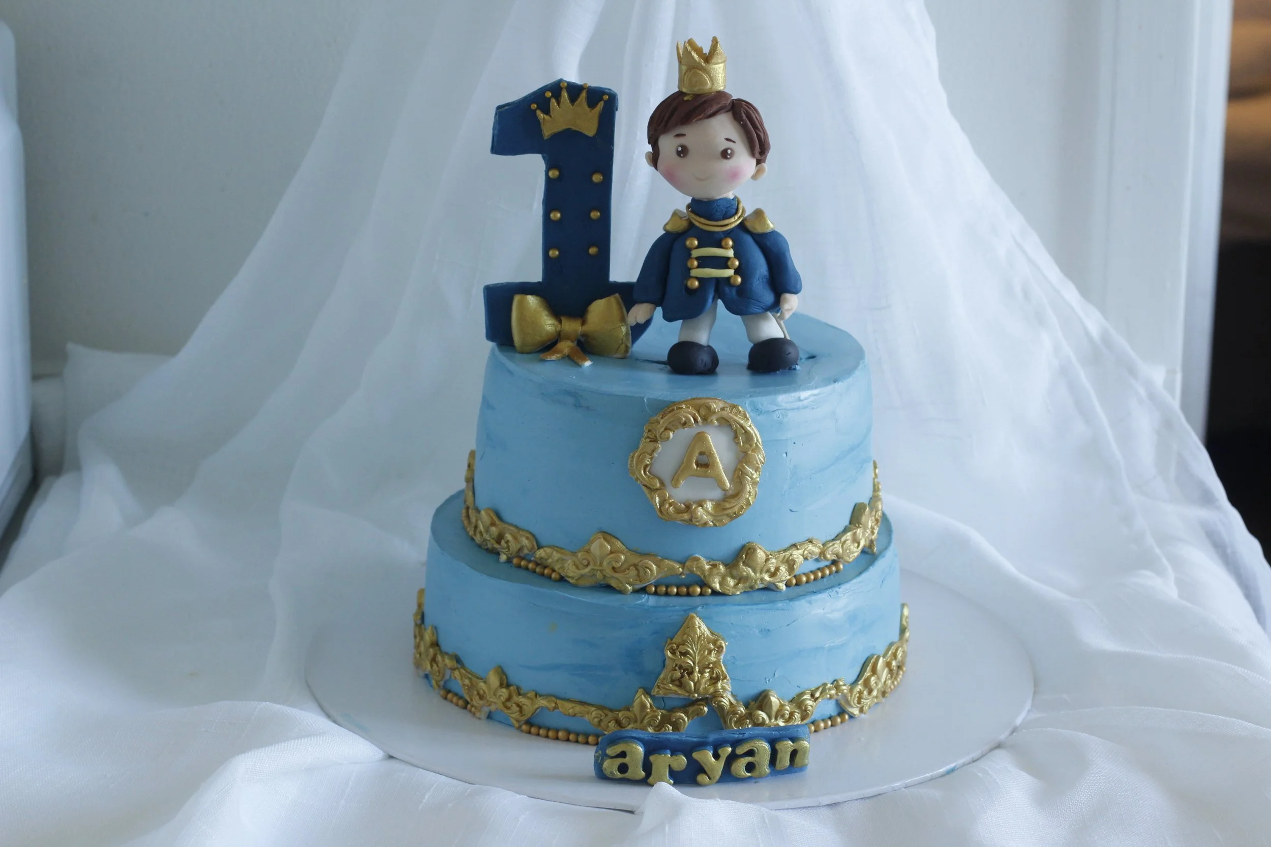 Two-tier blue birthday cake decorated with gold accents and a fondant figure of a boy prince on top, accompanied by a large number one candle and a gold and blue plaque with the name 'Aryan' at the bottom.