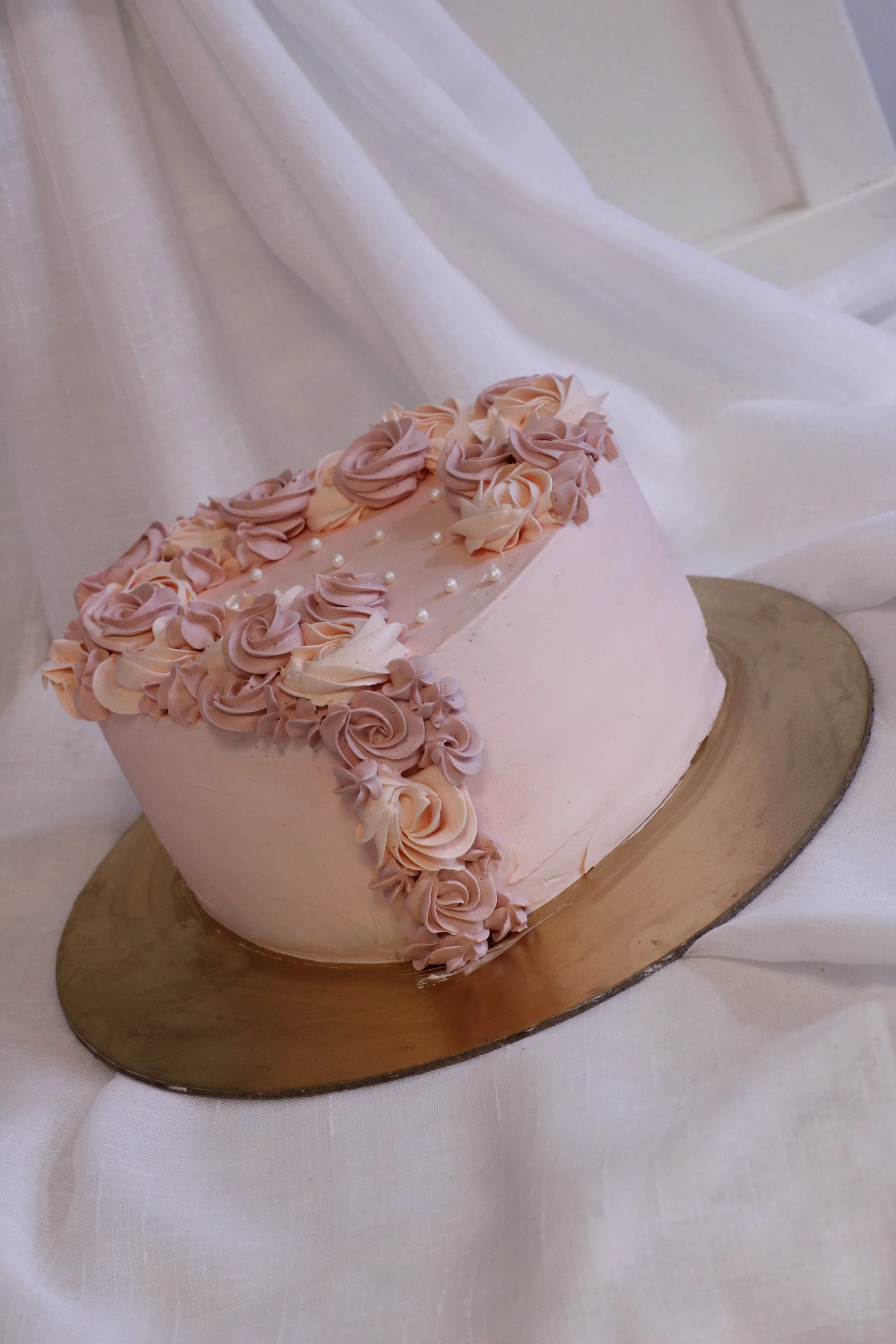 Pink heart-shaped cake decorated with pink and purple rosette frosting and small white pearls, placed on a gold cake board against a white fabric backdrop.