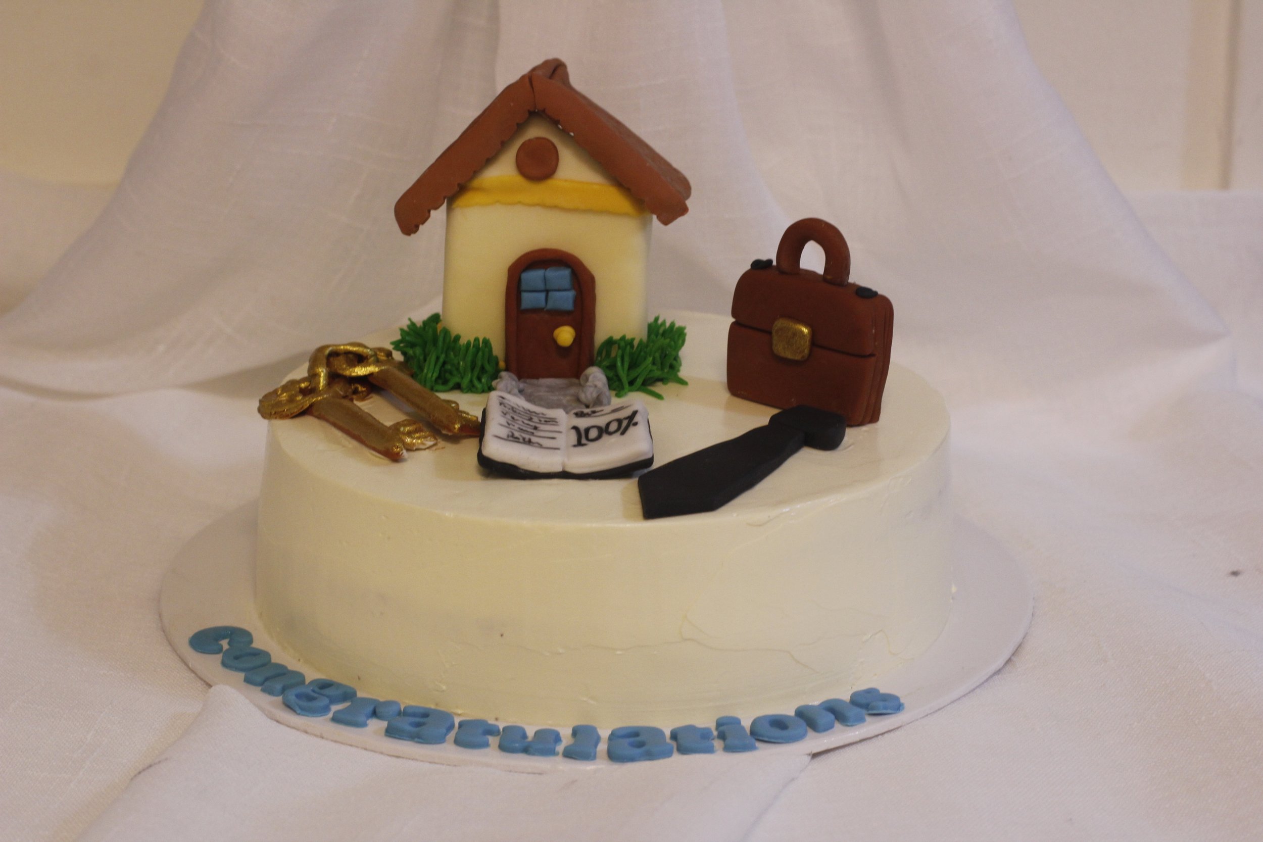 A round white-themed cake decorated with a miniature house, a briefcase, a black tie, gold keys, a newspaper, and a blue fondant message on the base that reads "Congratulations."