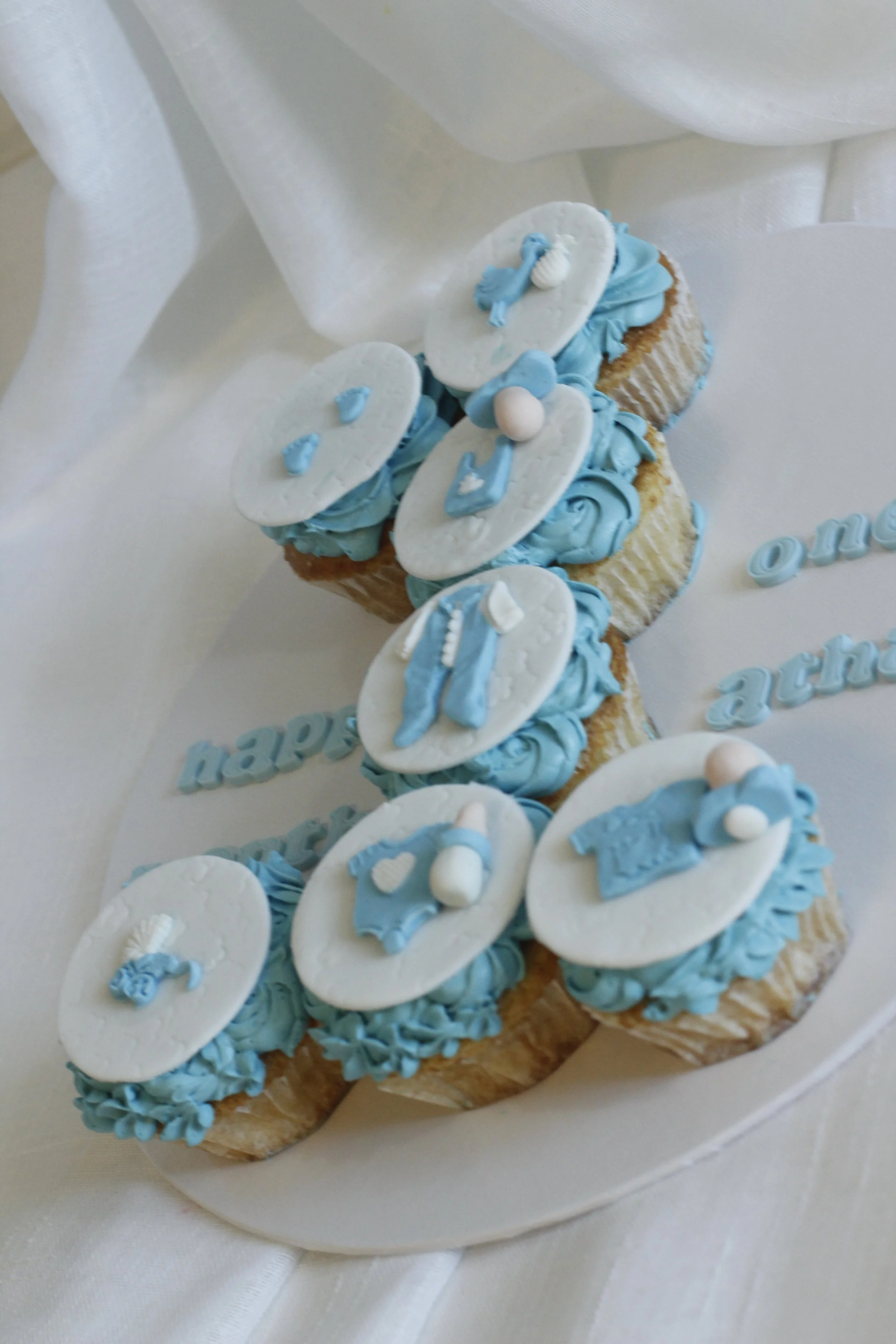 A decorative cake with blue and white baby-themed fondant toppers on top of the cupcakes and cake decoration. The cake is white with blue lettering.