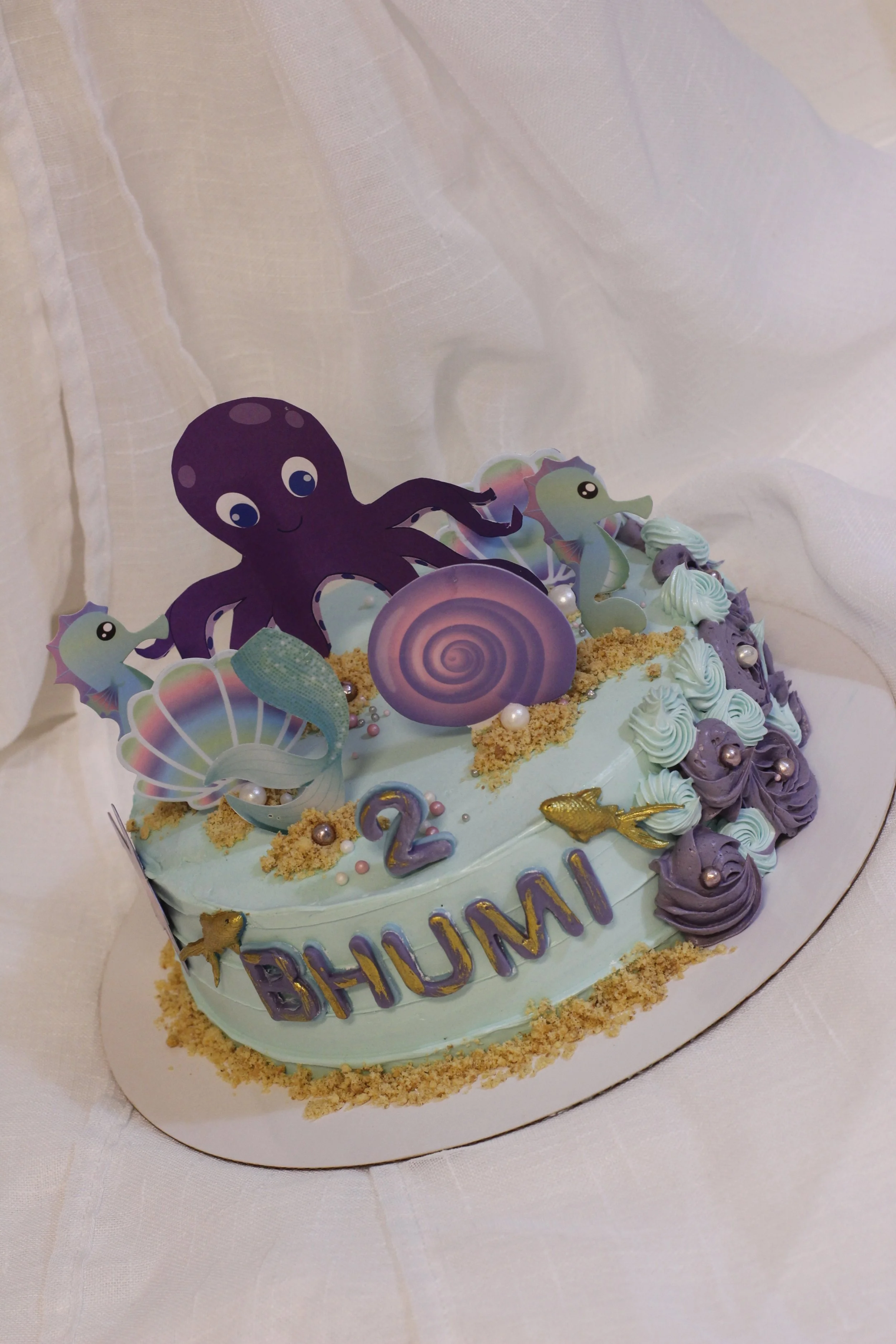 A birthday cake decorated with ocean-themed toppers including a cute purple octopus, seahorses, seashells, fish, and pearls. The cake has light blue frosting and says "BHUMI" in purple and yellow letters.