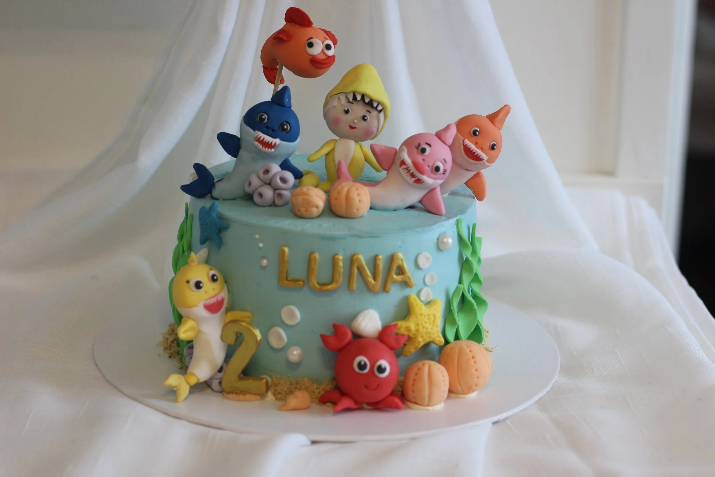 Birthday cake decorated with characters from the show Octonauts, featuring a red crab, yellow octopus, and other sea creatures, with the name 'LUNA' and the number 2 on it.