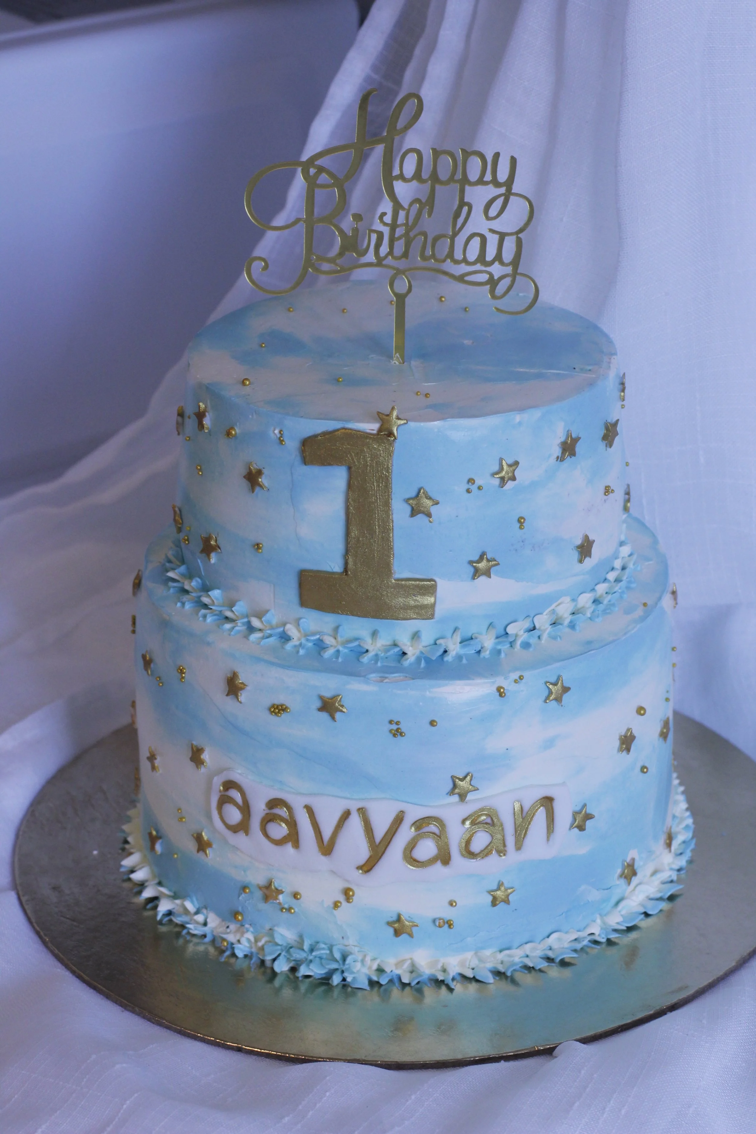 A two-tiered birthday cake with blue and white marbled icing, decorated with gold stars and a large gold number one. The cake has a gold birthday sign on top and the name 'davyän' written in gold letters on the front.