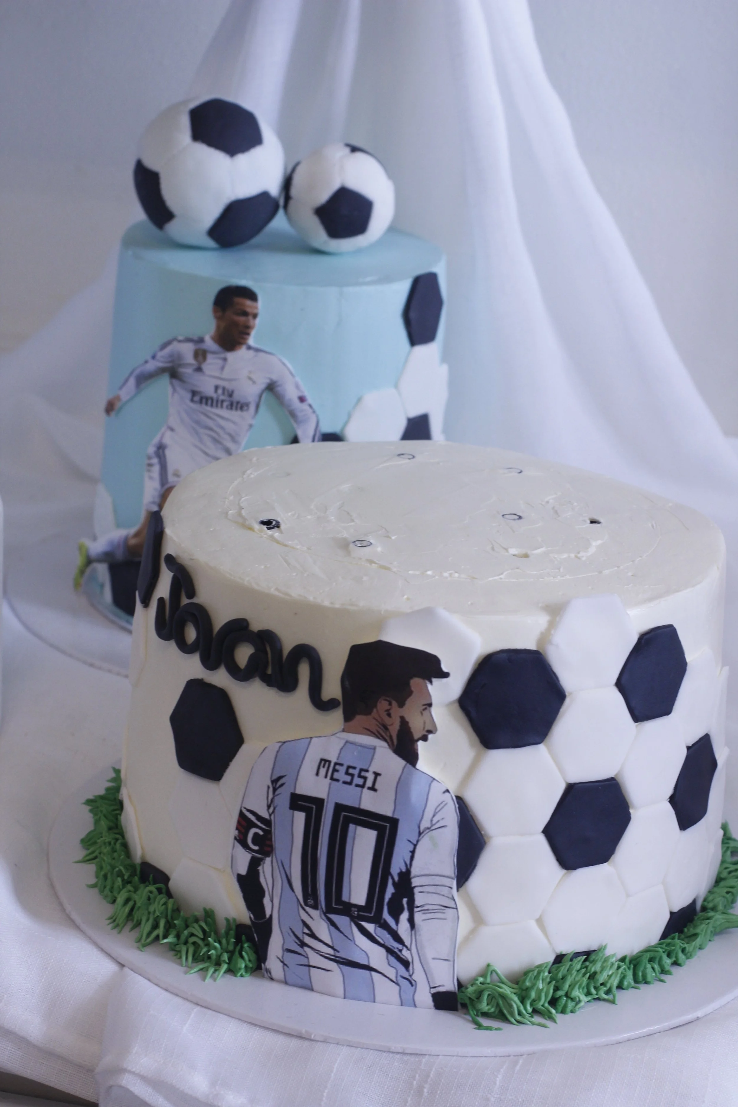 Two soccer-themed cakes decorated with soccer ball patterns, featuring images of Cristiano Ronaldo and Lionel Messi, with soccer ball plush toys on top.