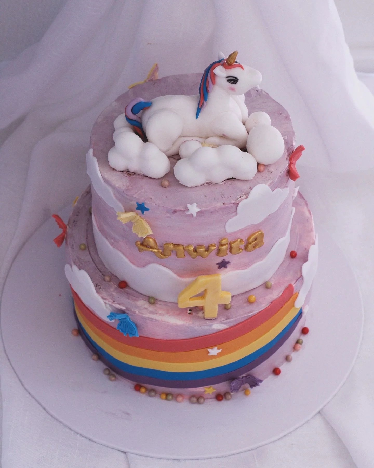 Pink and purple two-tiered birthday cake decorated with rainbow, clouds, stars, colorful butterflies, and small spheres, featuring a unicorn figure on top and the name 'Anvita' with the number 4.