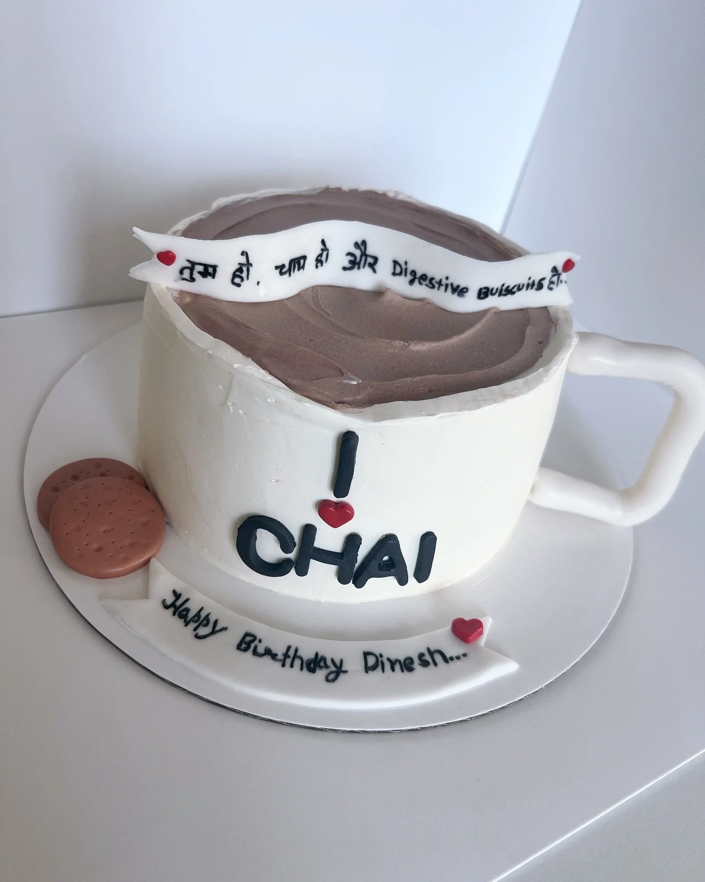 A birthday cake designed to look like a white mug filled with chocolate beverage, decorated with red hearts, cookies, and fondant message strips in Hindi and English. The cake has the message 'I love chai' and 'Happy Birthday Dinesh' written on it.