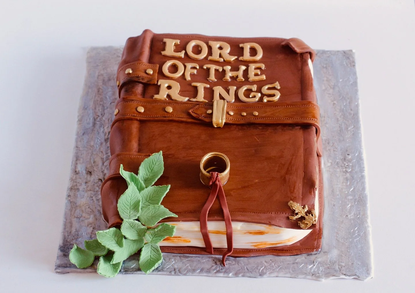 Cake shaped like a Bible with the words "LORD OF THE RINGS" on top, decorated with green leaves, gold accents, and a ring, placed on a textured silver base.