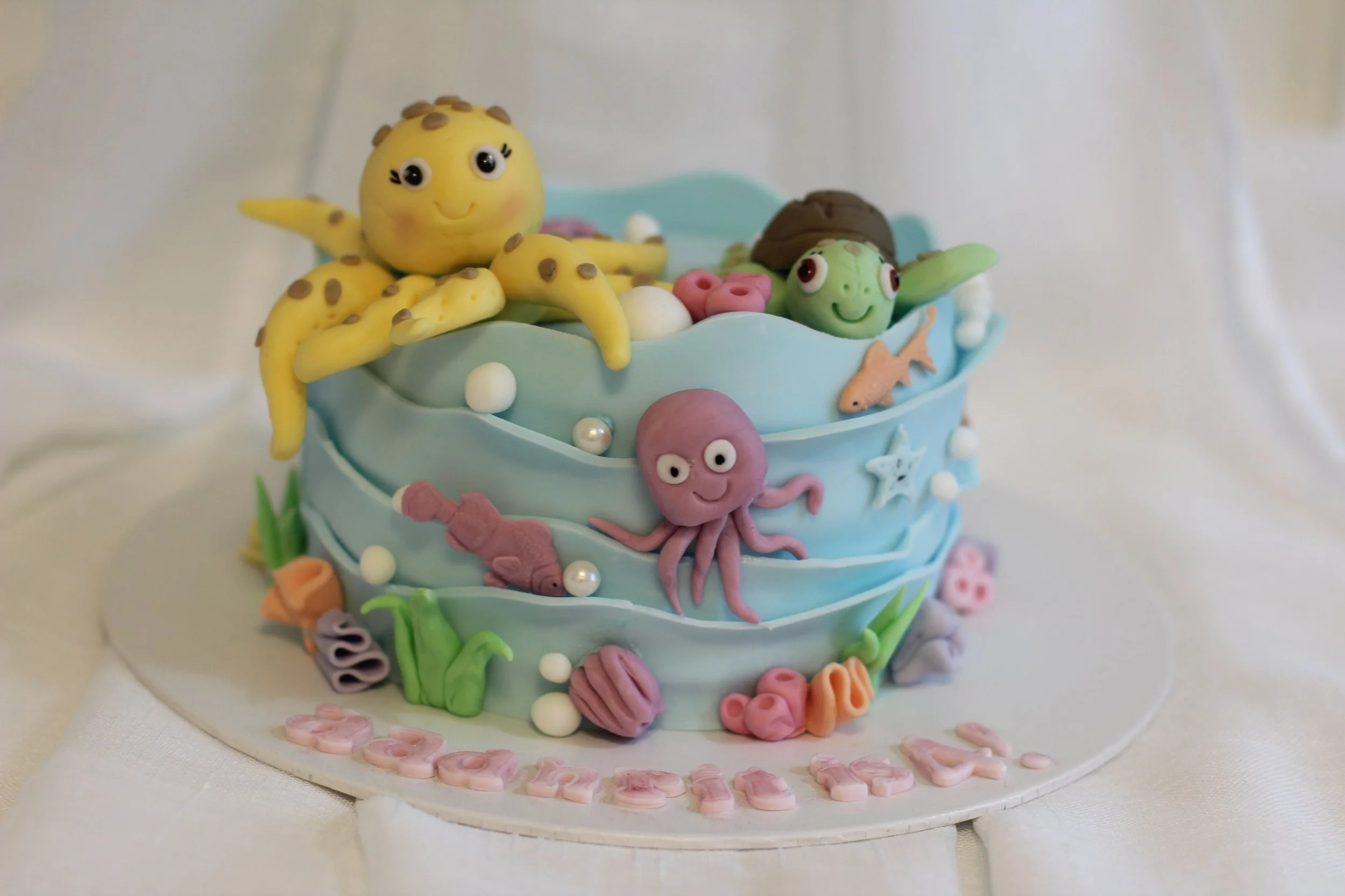 A two-tiered ocean-themed birthday cake decorated with fondant sea creatures including a yellow octopus, green turtle, pink octopus, purple starfish, and various seaweed, shells, and bubbles, with pink and white flowers on the base and pink lettering