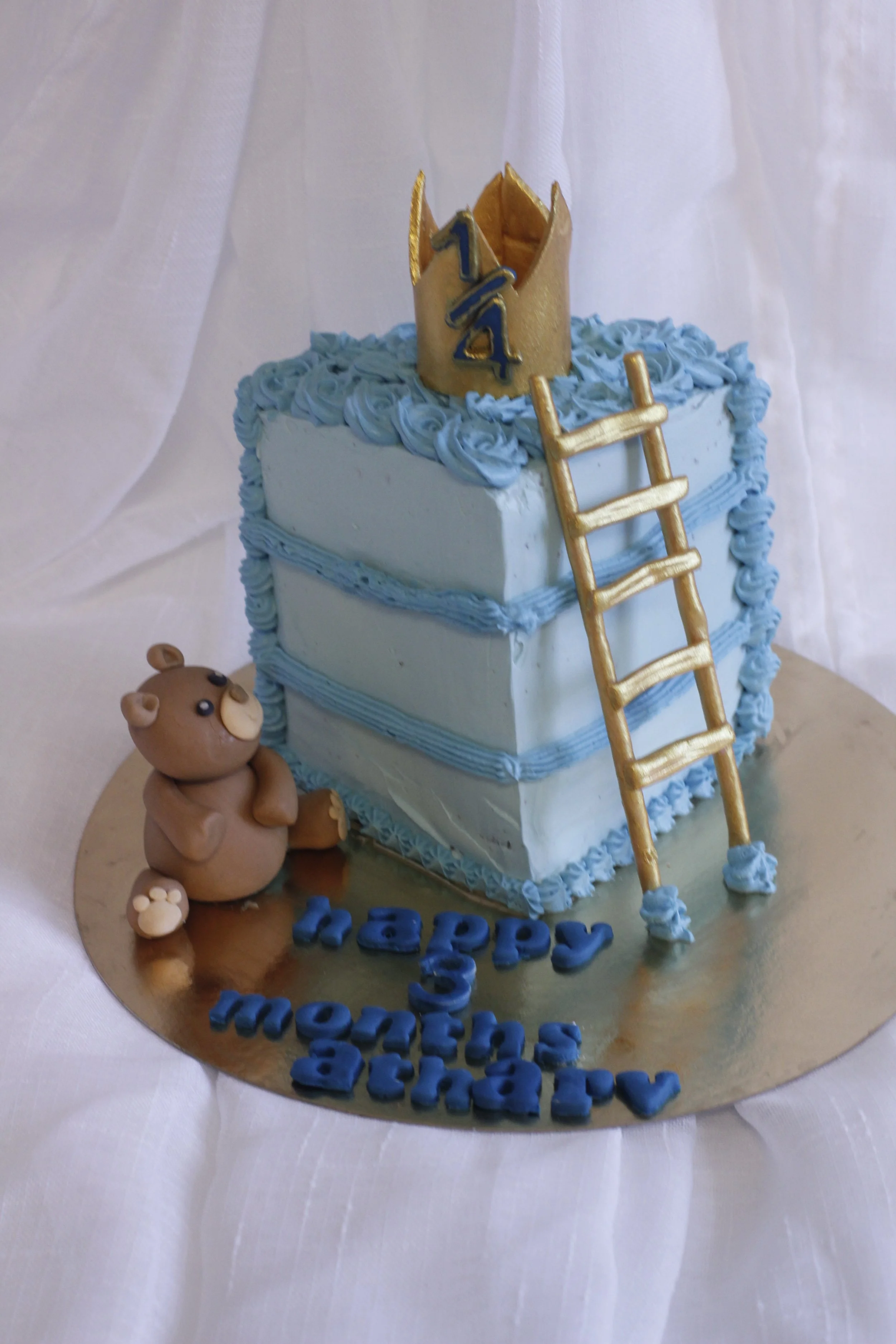 A birthday cake with light blue frosting, a small bear figurine, and a gold crown with the number 14 on top. The cake has a gold ladder leaning against it. The message on the cake reads "happy momma anniversary" in blue letters.