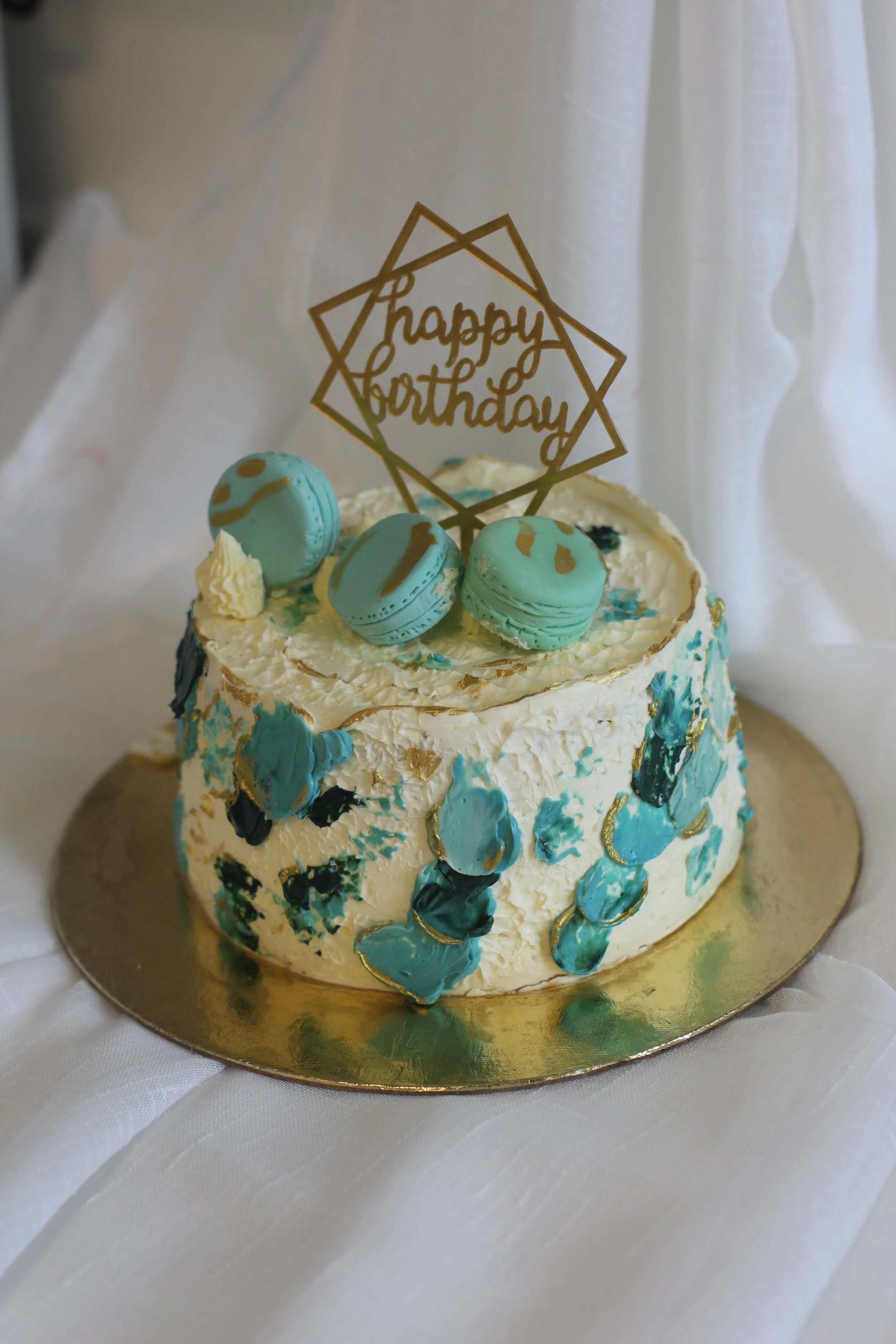 A birthday cake decorated with blue macarons, white and blue frosting, and a "happy birthday" topper, sitting on a gold cake board and surrounded by white drapery.