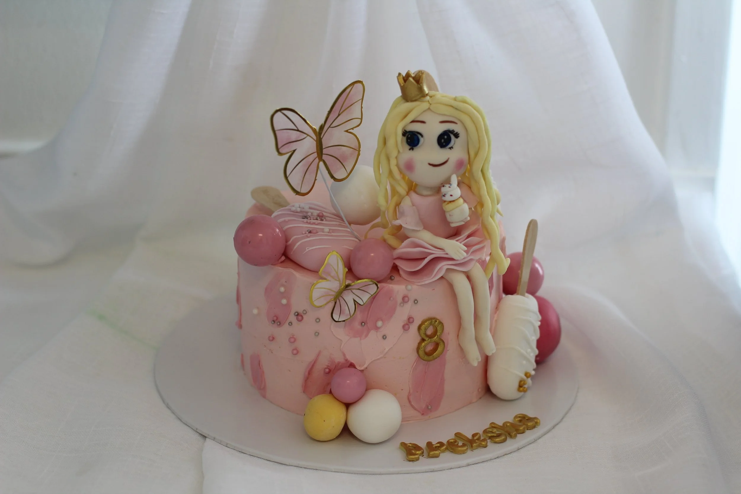 Pink birthday cake with a princess figurine on top, decorated with butterflies, colorful balls, and a white popsicle, with an '8' number and the name 'Rebekah' in gold letters.