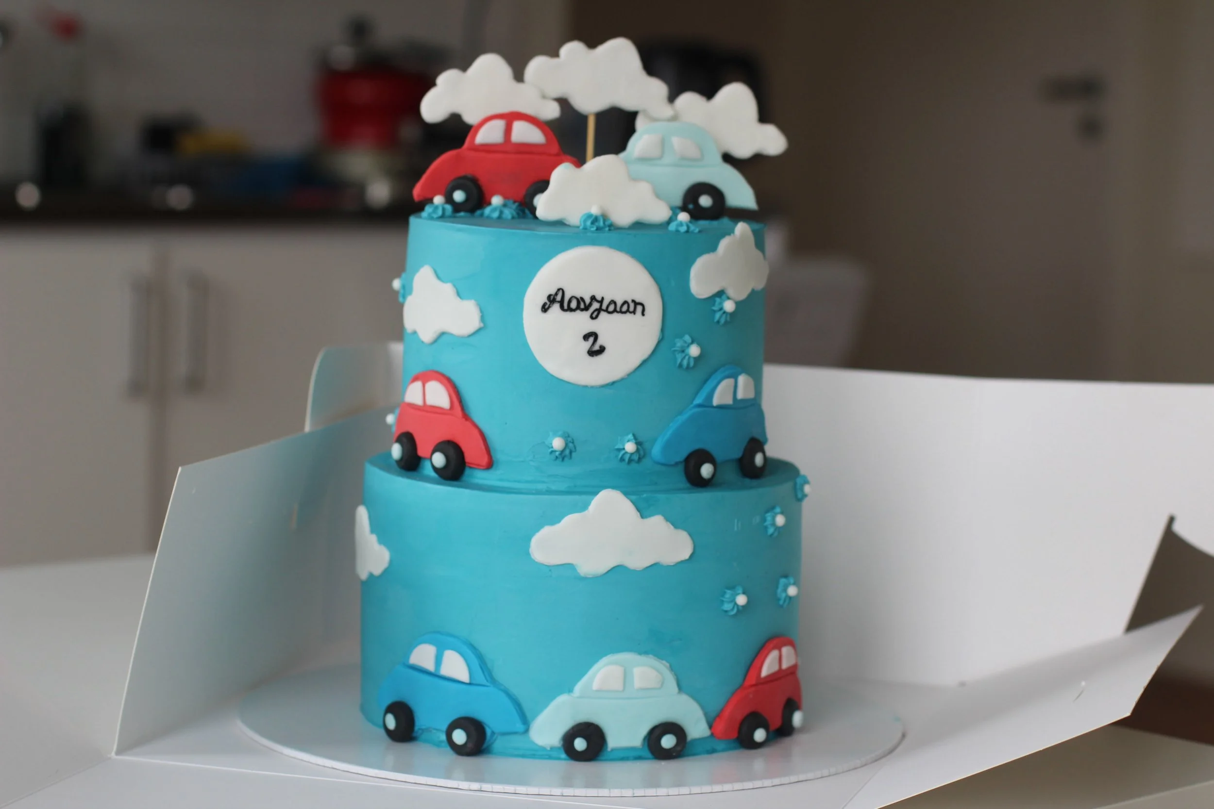Blue two-tiered birthday cake decorated with colorful fondant cars, white fondant clouds, and small blue and white flowers, with a white circular fondant plaque on top inscribed with 'Aayaan 2'.
