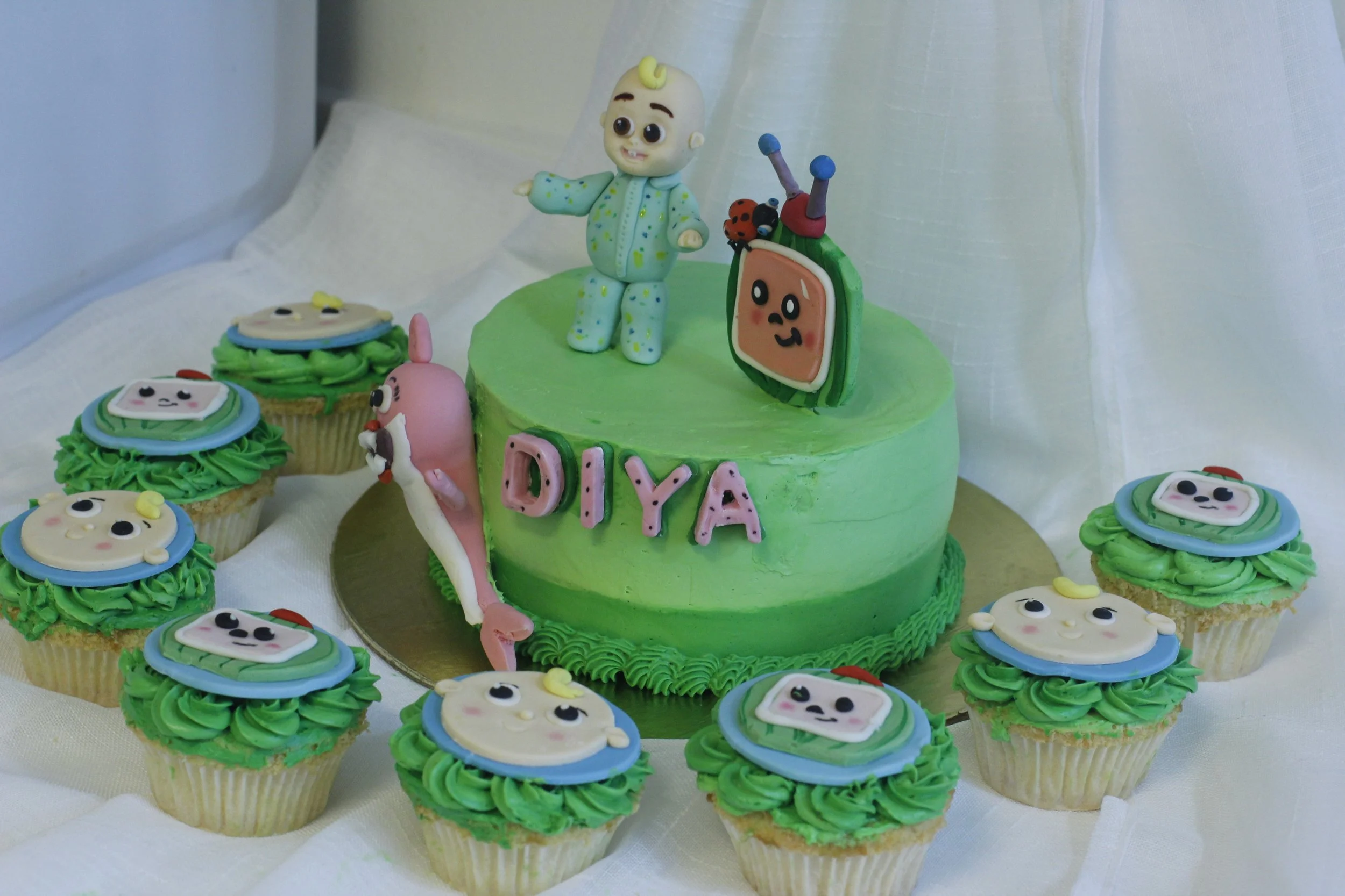 A birthday cake with green frosting and a toy of a baby with a yellow curl on top, decorated with the name 'Diya', surrounded by cupcakes with character faces on green frosting.