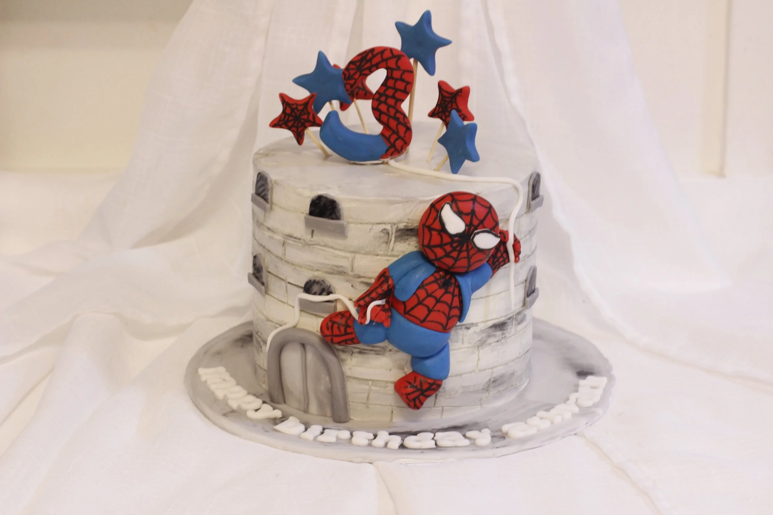 Spider-Man themed birthday cake with a Spider-Man figurine climbing a wall and cake toppers of stars and webs, decorated in red, blue, and grey colors.