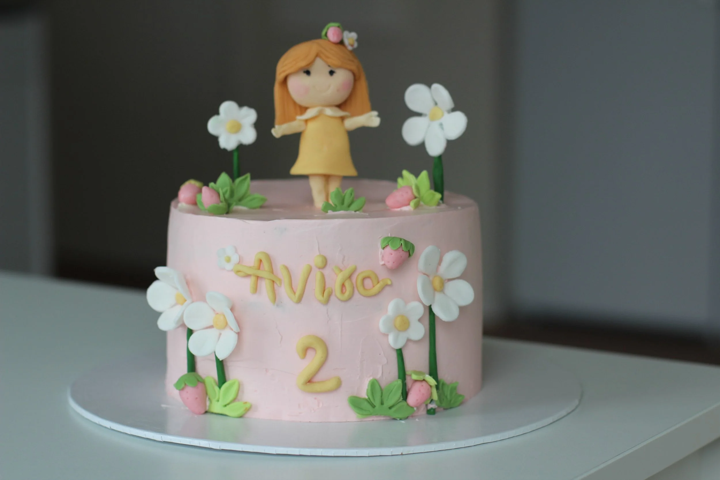 A two-tier pink birthday cake decorated with white flowers, strawberries, and a fondant figure of a girl with orange hair in a yellow dress on top. The cake has the name "Aviça" and the number "2" written on it.