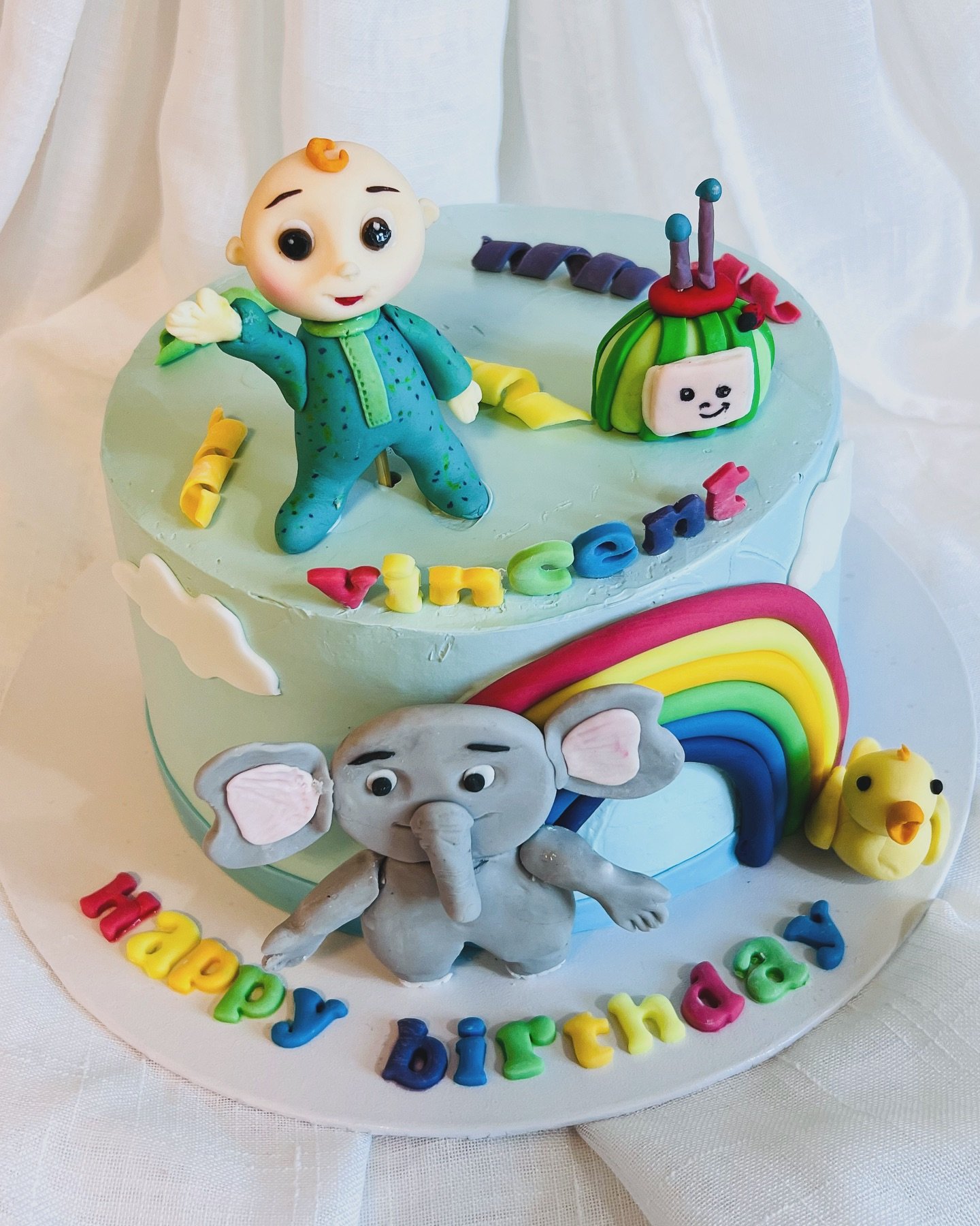Two-tier birthday cake decorated with pastel blue icing, featuring figurines of a baby in a blue outfit, an elephant, a yellow duck, a rainbow, a toy train, a green watermelon, and colorful letters spelling 'Happy Birthday', with a white cloth backgr