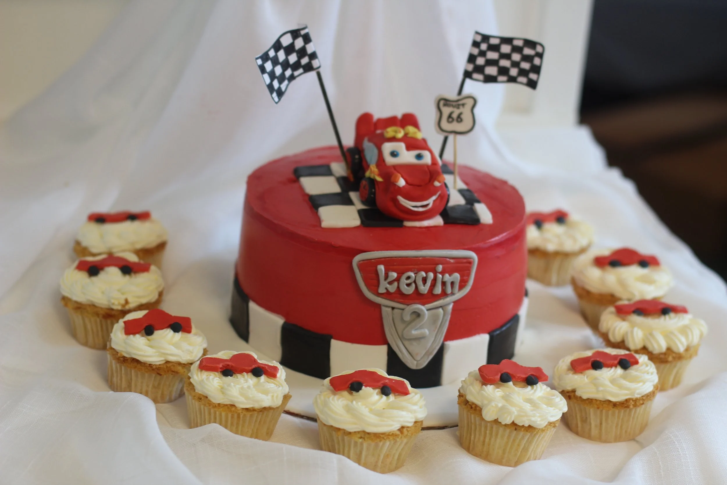 A birthday cake themed around racing with a red car figurine on top, surrounded by ten cupcakes decorated with racing car motifs, on a white cloth background.