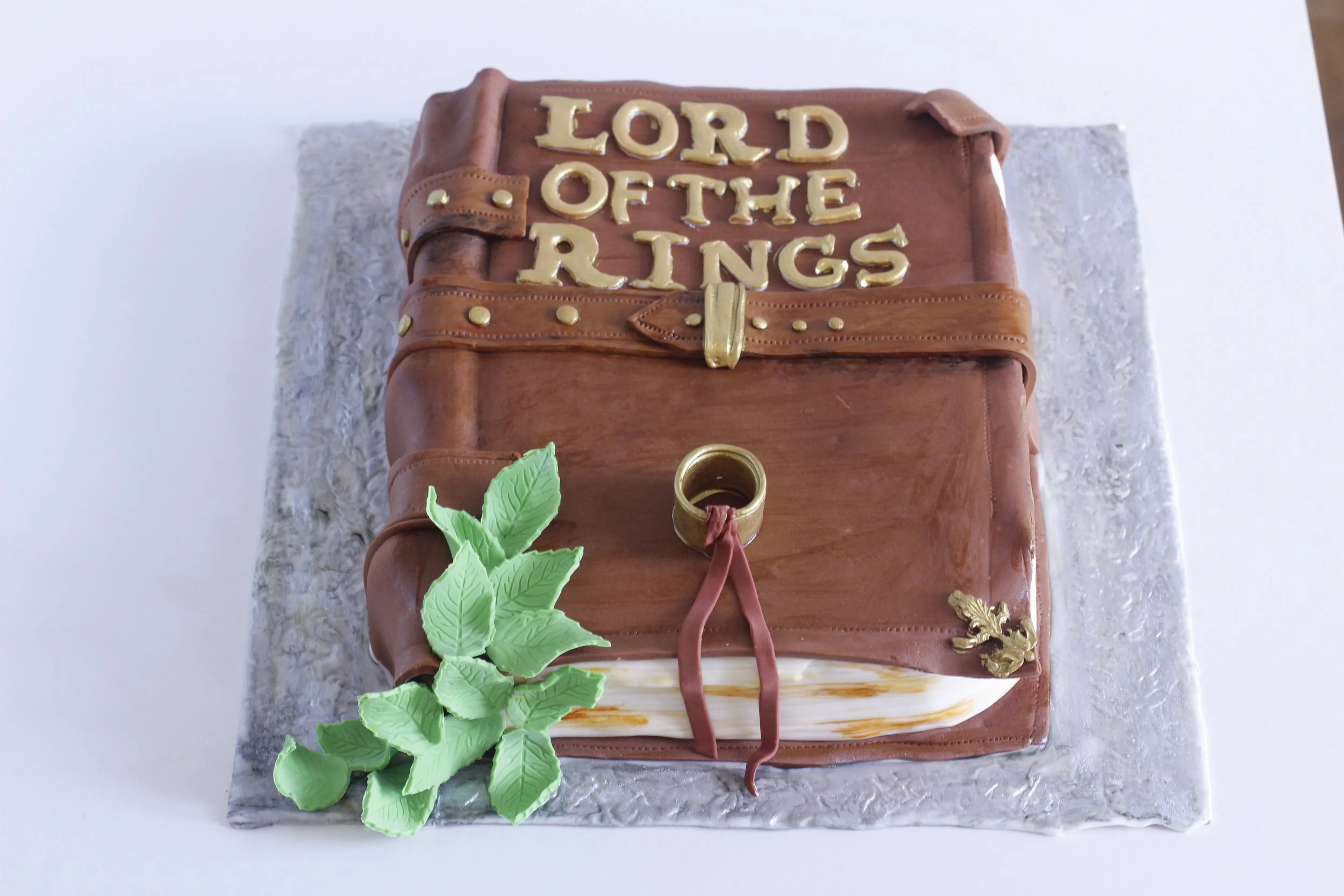 A cake decorated as a leather-bound book, themed after The Lord of the Rings, with gold letters spelling 'Lord of the Rings', green leaves, and a scroll design.