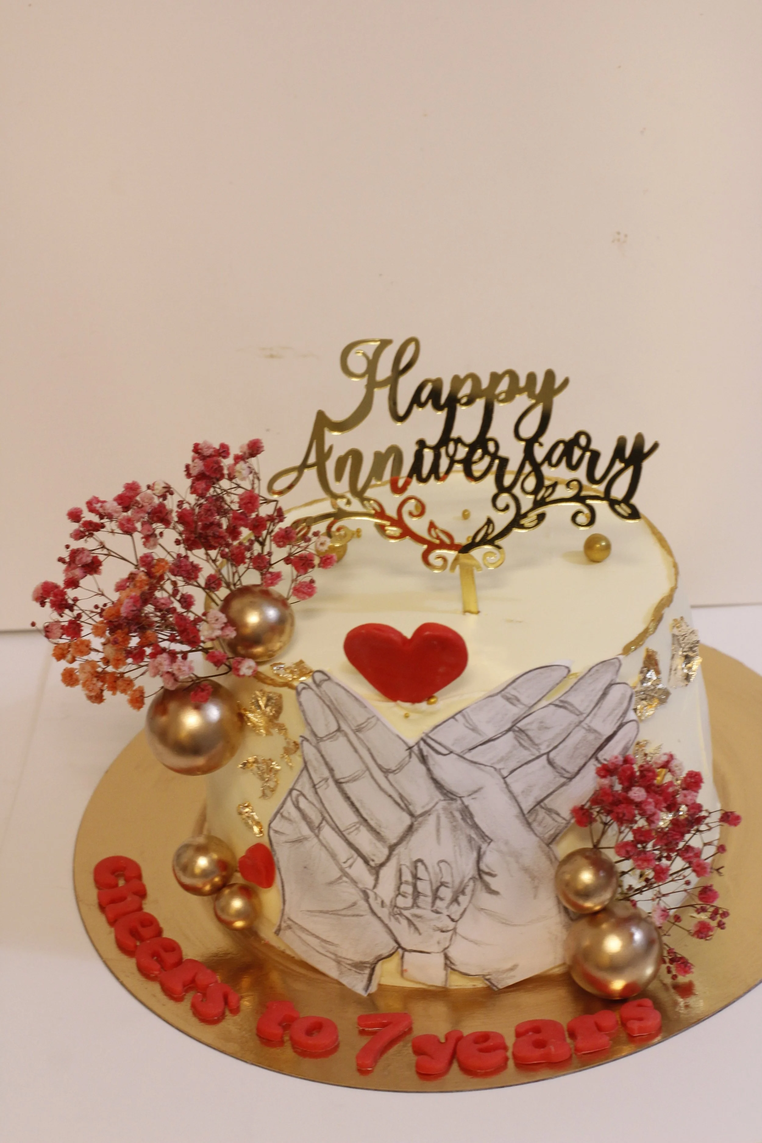 A decorated birthday cake with a drawing of two hands, one small and one large, touching, surrounded by pink flowers, gold accents, and heart decorations. The cake has a topper that says 'Happy Anniversary' and red text reading 'Cheers to 7 years'.