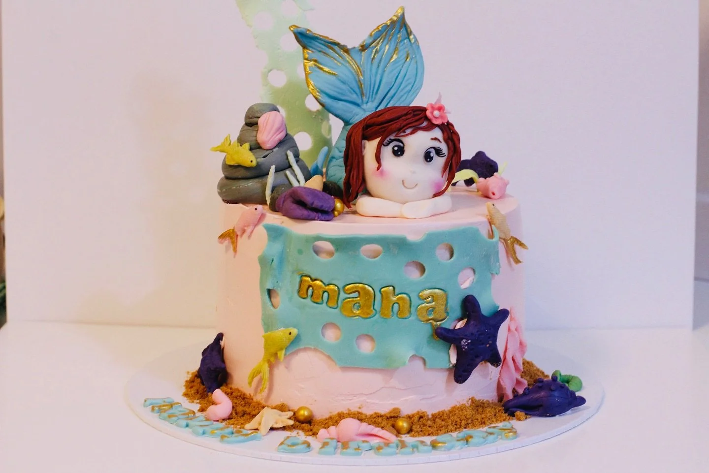 A mermaid-themed birthday cake with pink frosting, featuring a fondant mermaid figure with red hair, a large blue tail, and ocean animals like starfish, sea shells, and fish, with gold accents and the name 'Maha' written in gold on the front.