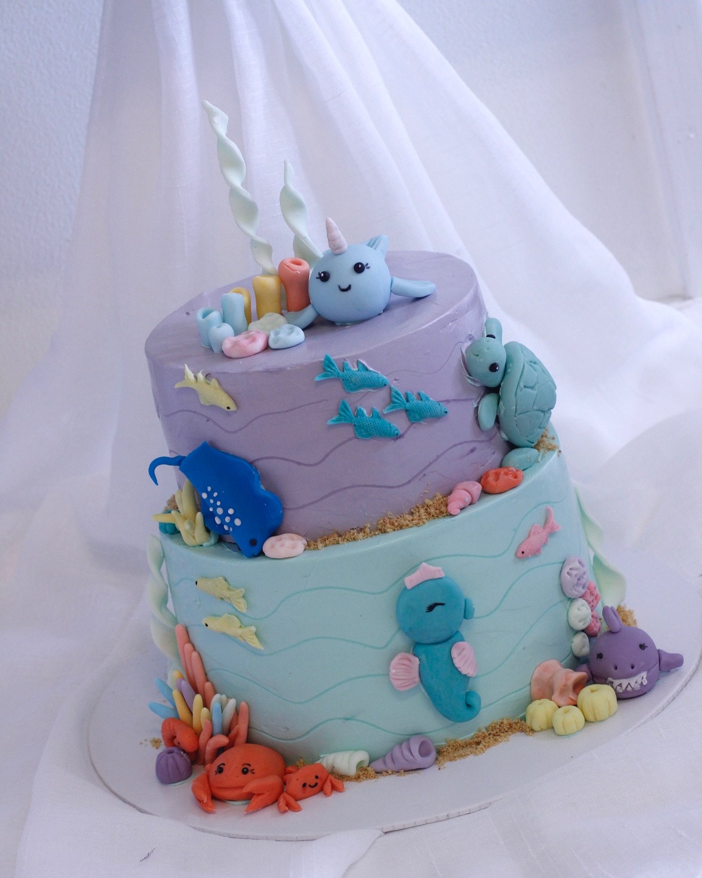 Colorful two-tiered ocean-themed cake with fondant sea creatures like a unicorn, turtles, fish, seahorses, sharks, crabs, and coral decorations.