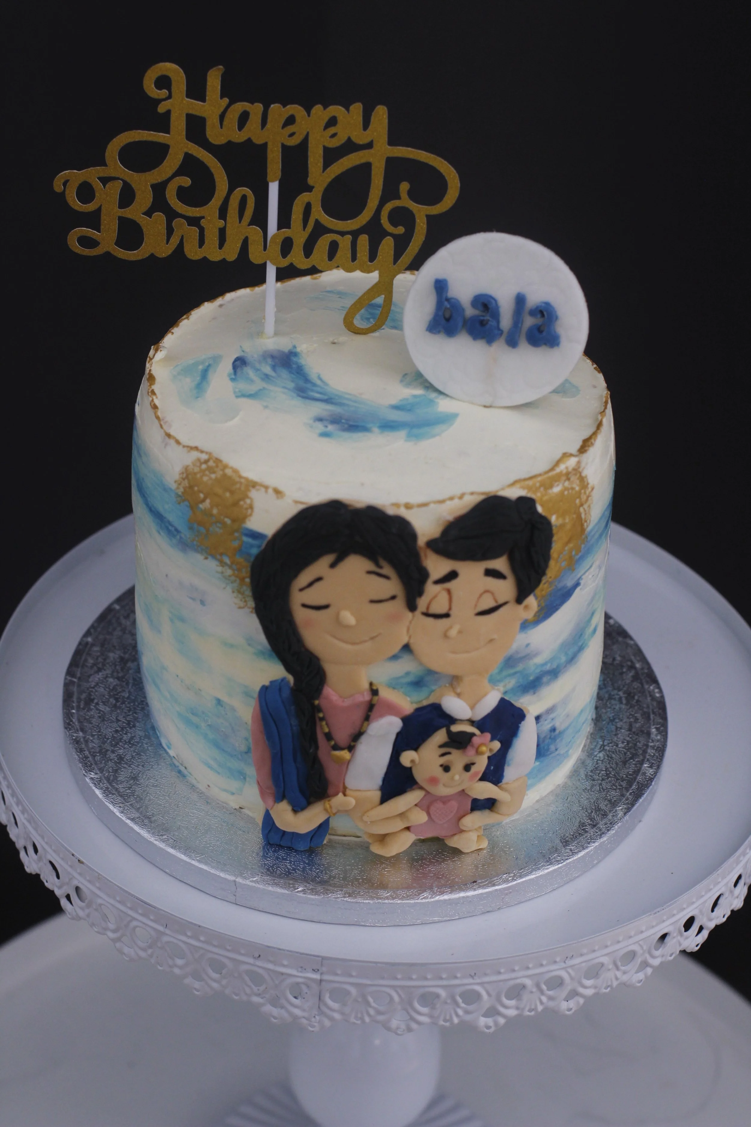 Decorative birthday cake with a blue and white wave pattern, topped with a gold 'Happy Birthday' cake topper, a white circular topper with 'bala' in blue, and a fondant family figurine of a smiling man, woman, and baby, on a white cake stand with lac