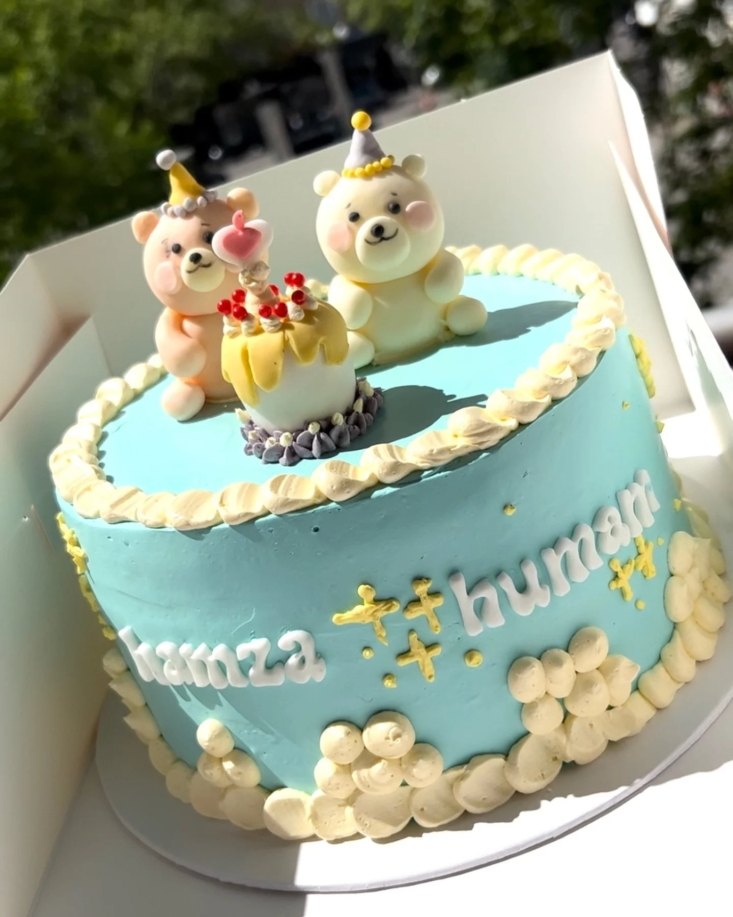 A blue birthday cake decorated with white frosting, featuring two bear figures wearing party hats and a small cake with candles on top. The cake has the text 'hamza + khumar' written on the side.