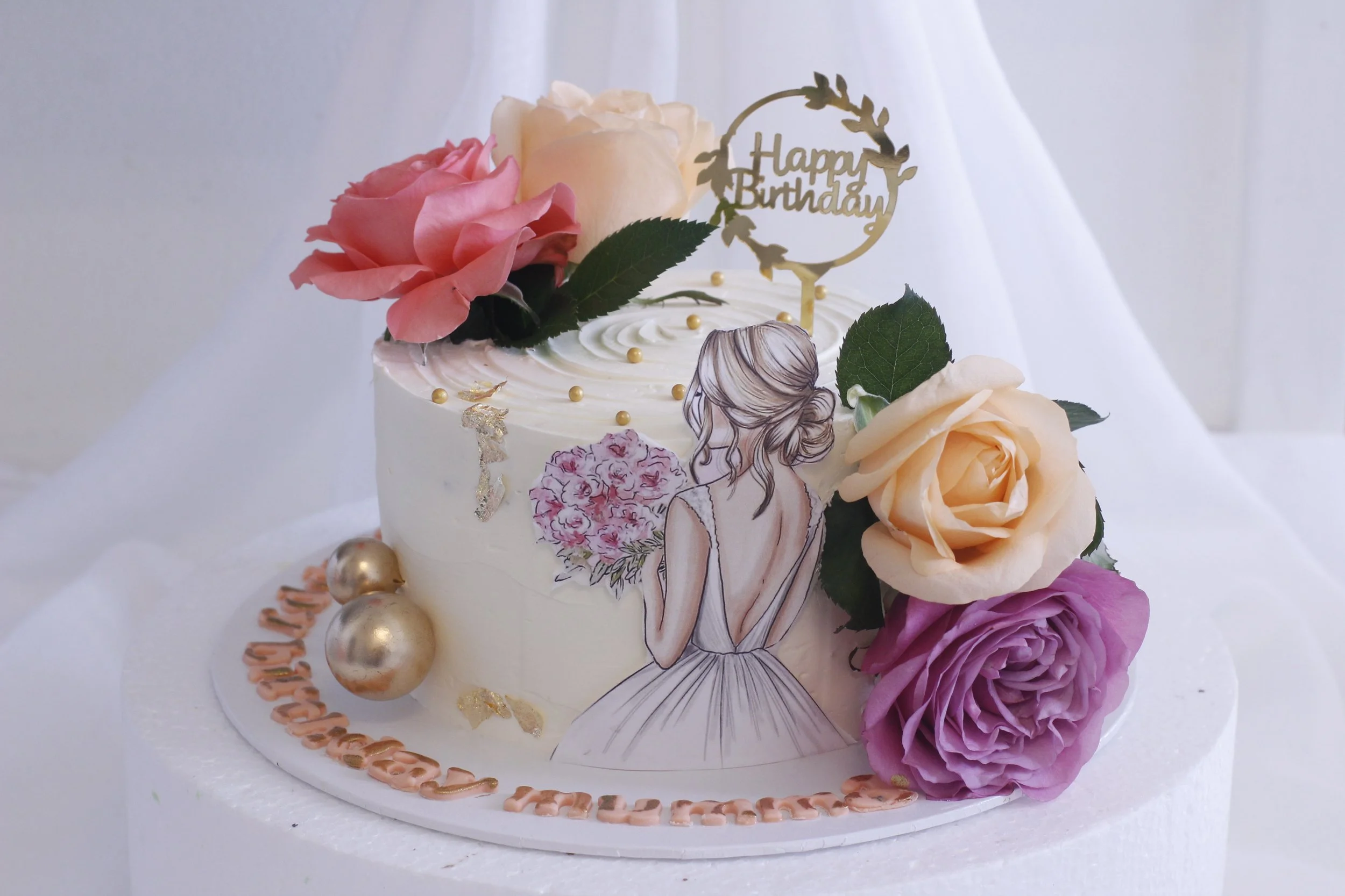 A decorated birthday cake with fresh roses, gold accents, and an illustration of a woman holding a bouquet. The cake has a 'Happy Birthday' topper and is adorned with gold and pearl decorations.