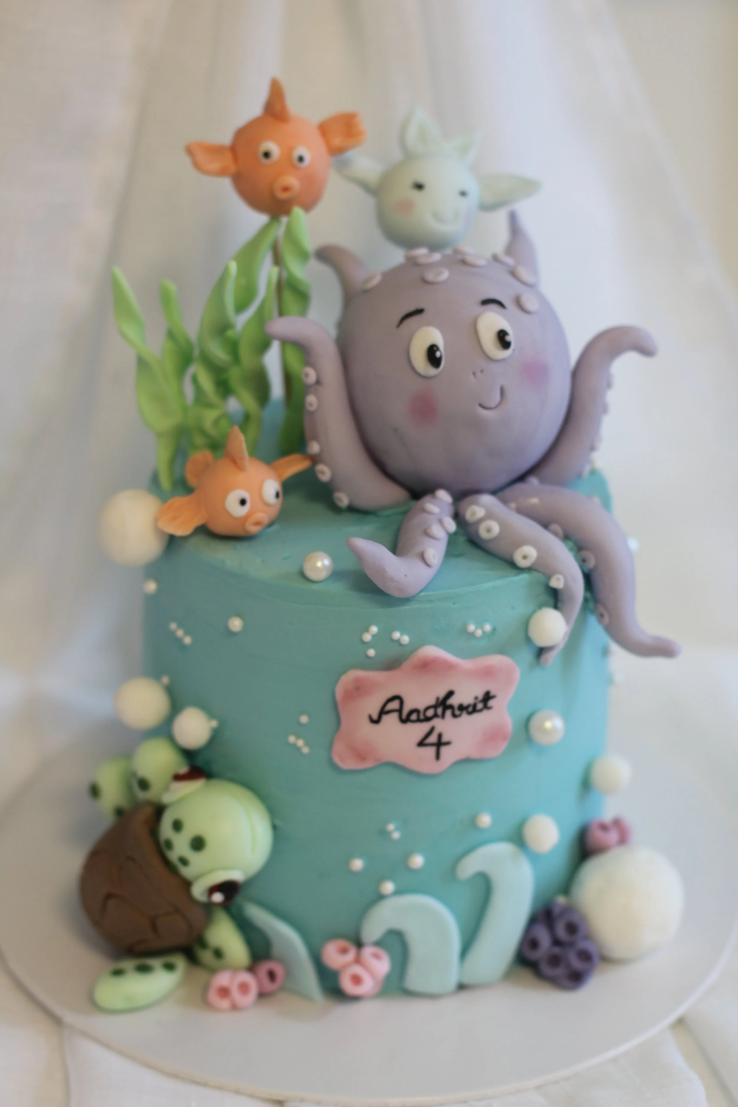 A birthday cake decorated with aquatic-themed fondant figures including an octopus, fish, and seaweed. The cake has a pink plaque that reads 'Aadhvit 4' and is adorned with pearls and pastel-colored embellishments.