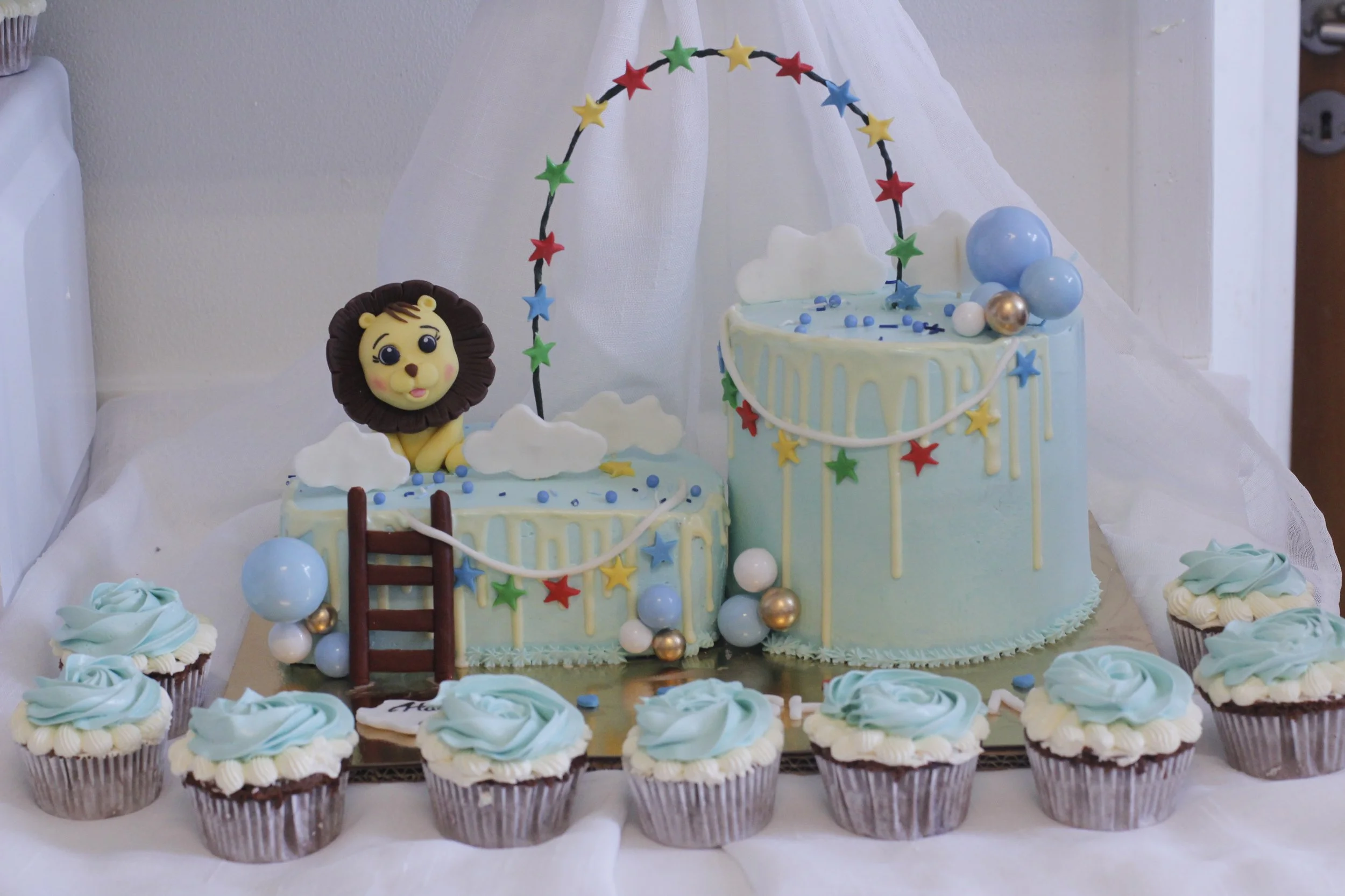 A set of two light blue birthday cakes decorated with clouds, stars, and balloons, with a lion figure on top, surrounded by ten cupcakes with blue and white frosting.