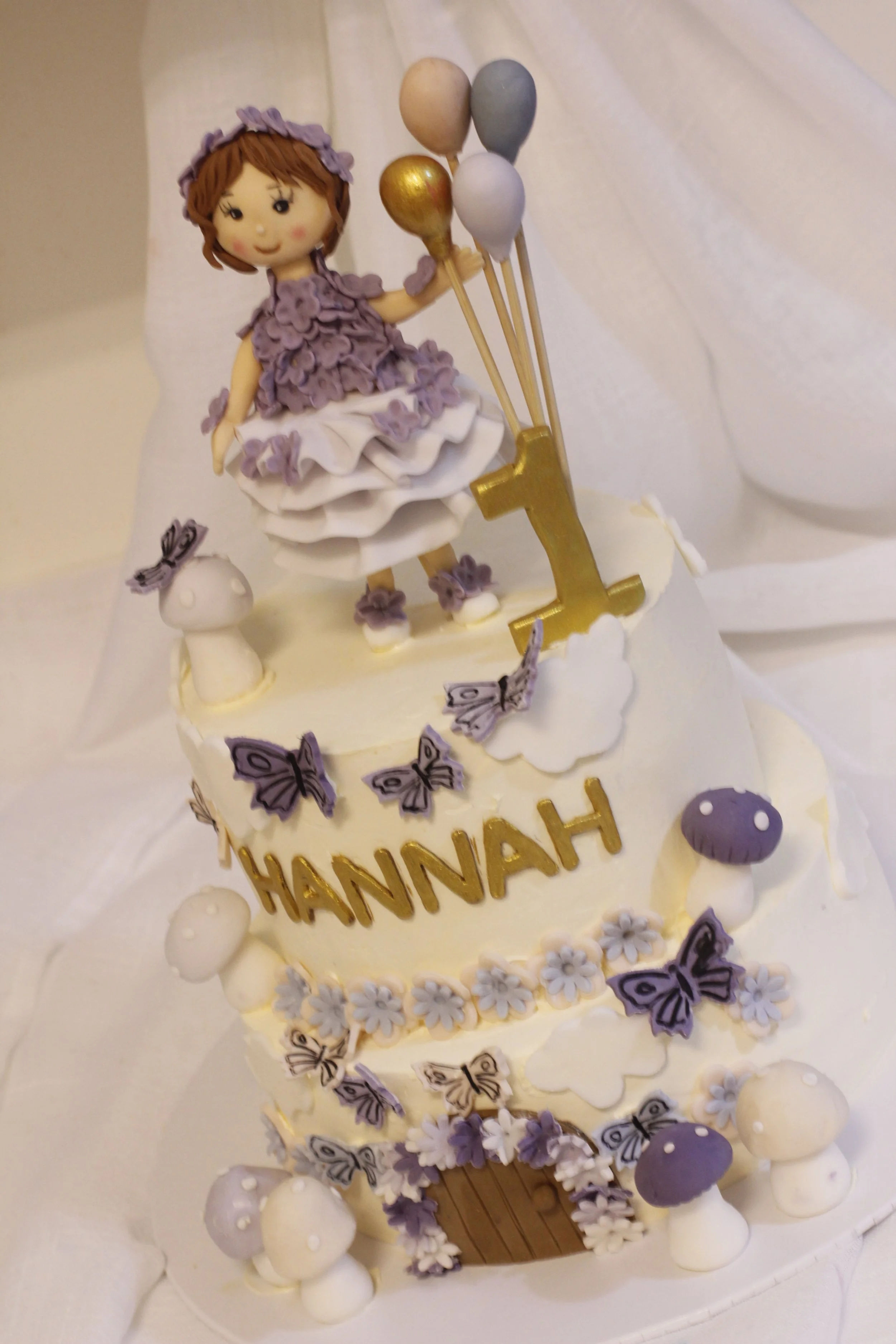 A decorated birthday cake with a purple and white theme. The cake has a figure of a girl in a purple dress and flowers, a gold number one, and balloons on top. The name 'Hannah' is written on the side of the cake with purple butterfly and mushroom de
