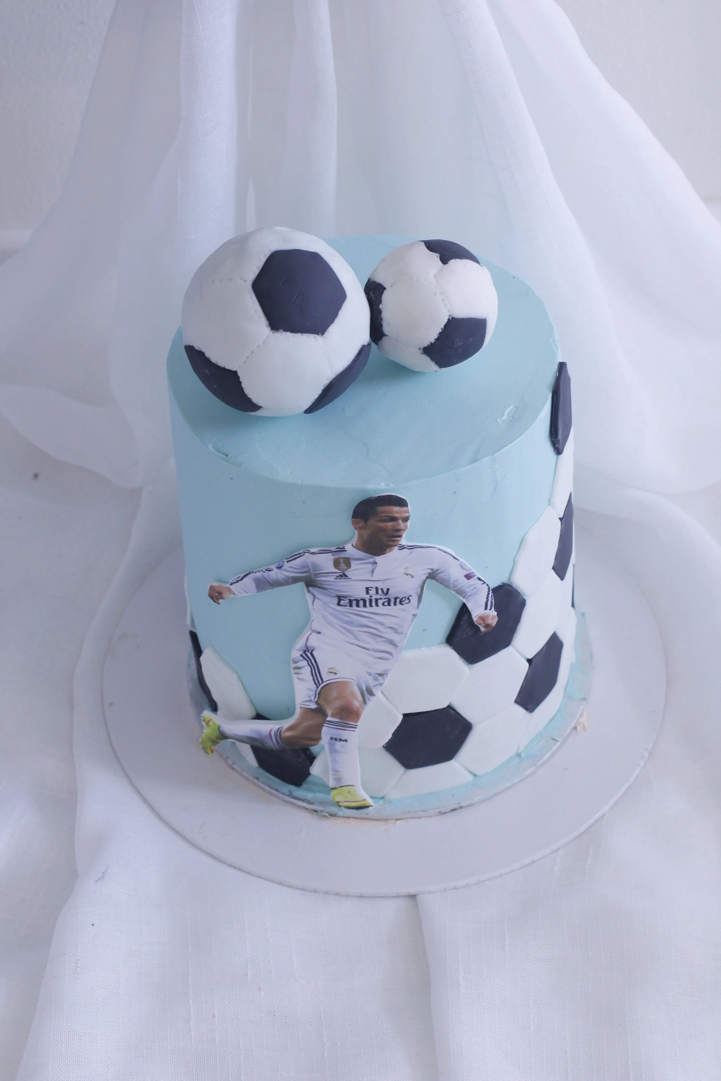 A birthday cake decorated with football-themed elements including soccer ball toppers, a Real Madrid player cutout, and black and white soccer ball patterns.