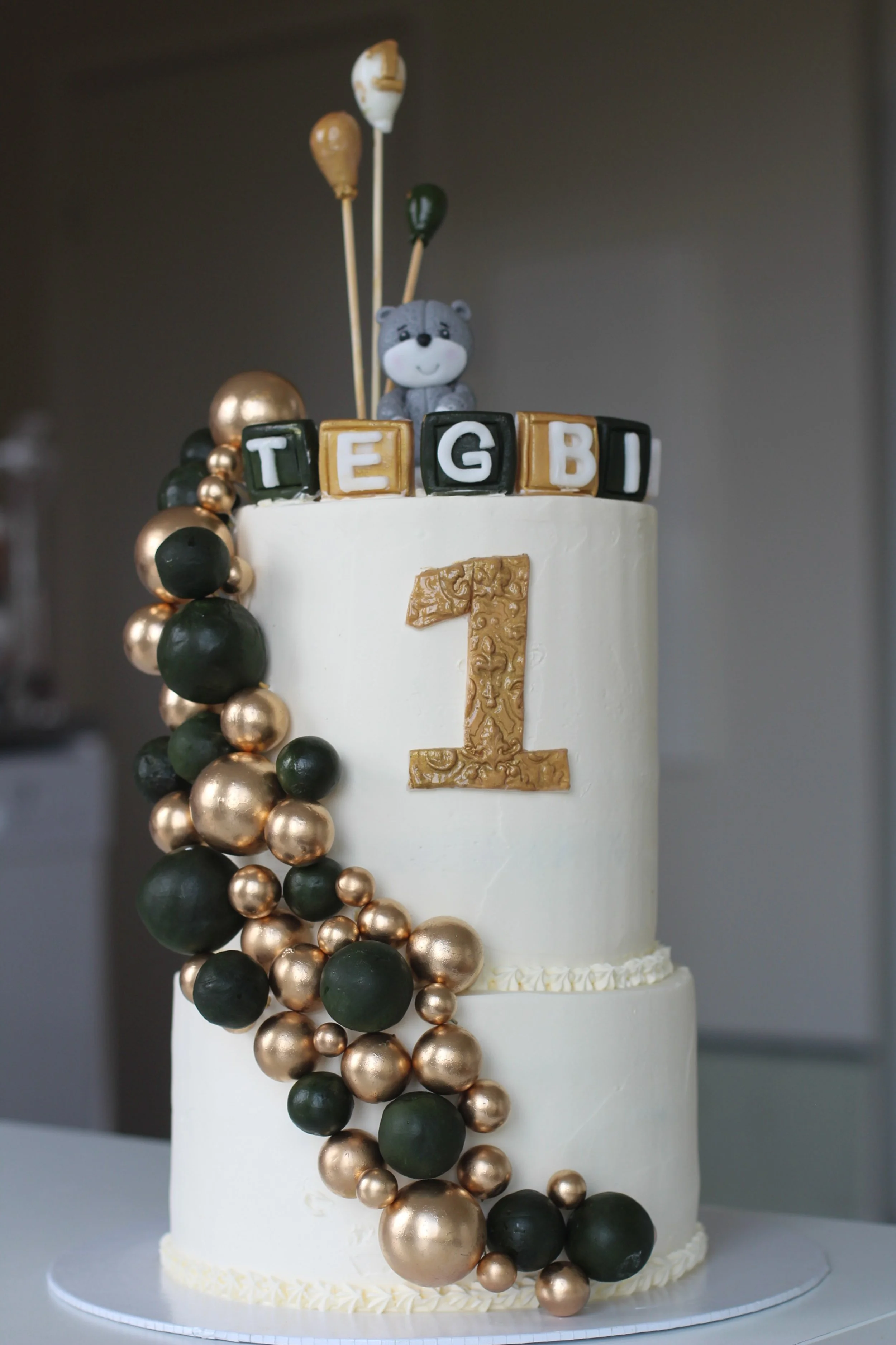 A two-tier white birthday cake decorated with gold and black beads, with a large gold number one on the front, topped with a small teddy bear and black block letters spelling TEG BI.