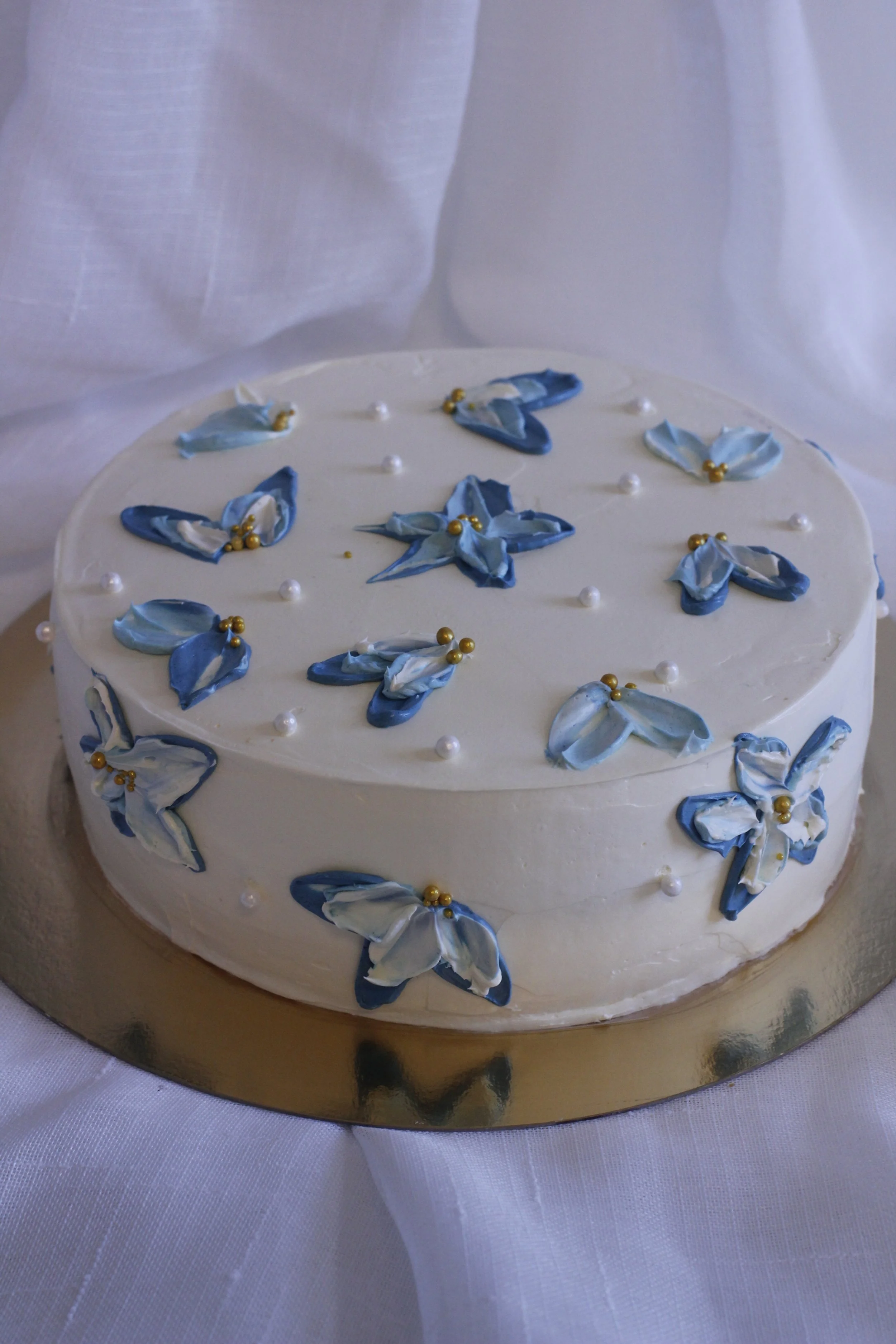 A round white cake decorated with blue and white flower-shaped icing and small golden beads on top, sitting on a gold cake board.