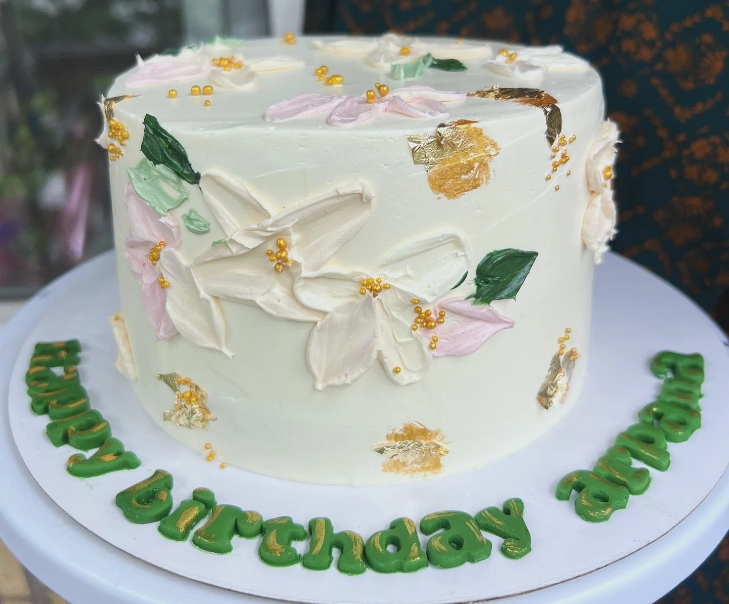 White birthday cake decorated with pink, green, and gold accents and the message "Happy Birthday" in green letters with gold highlights. The cake has floral and leaf decorations made of icing and gold foil.